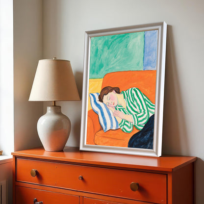 A framed painting of a woman sleeping on an orange sofa rests on a bright orange dresser next to a large ceramic lamp. The painting has a white frame.