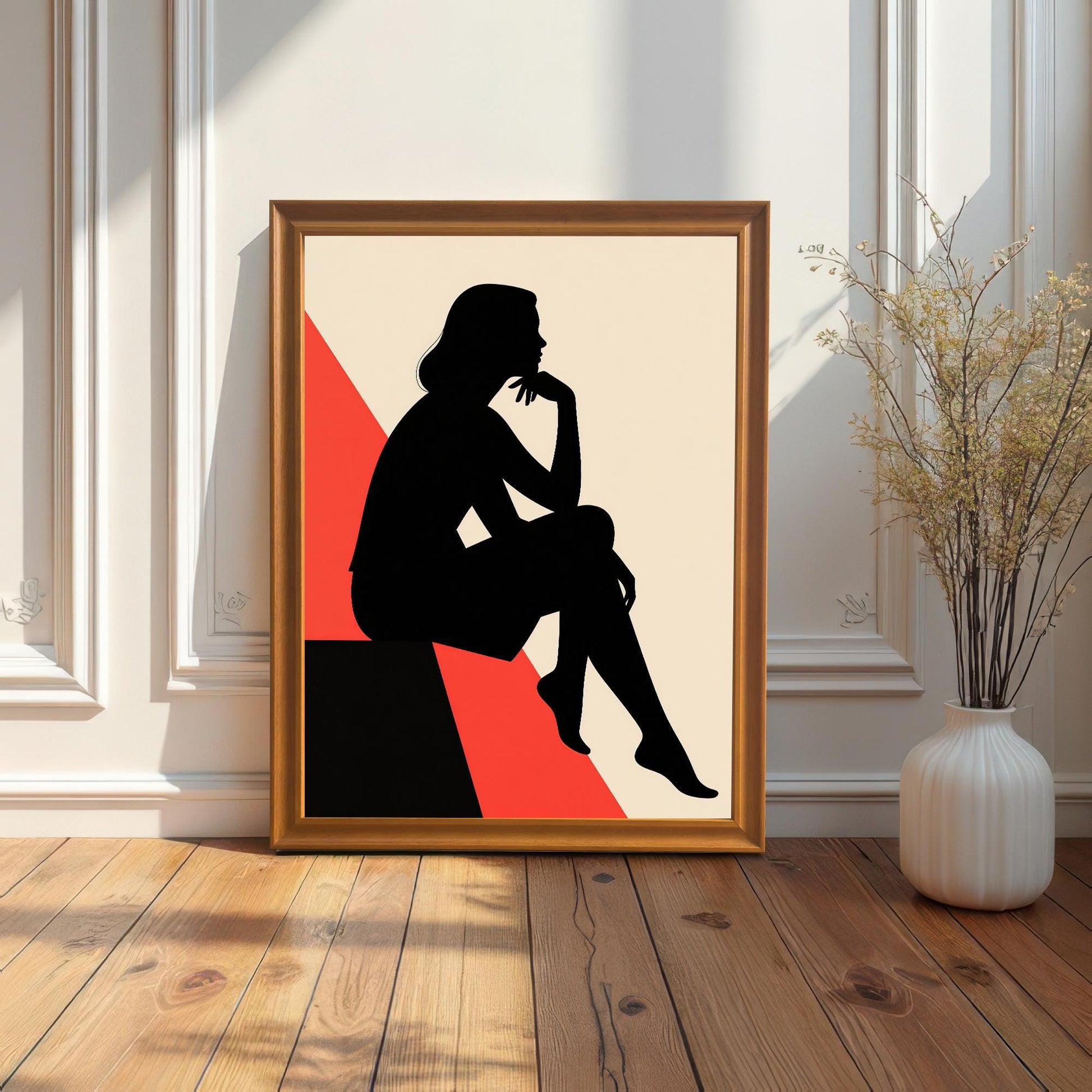 A framed art print with a gold frame is shown leaning against a white paneled wall. The frame sits on a light wood floor next to a white vase with dried plants.