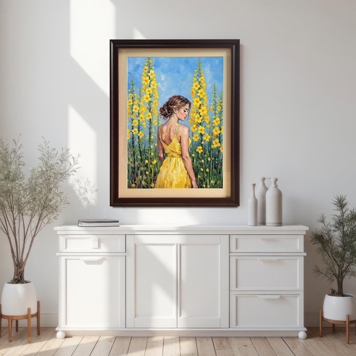 An oil painting of a woman in a yellow dress standing among tall yellow flowers, framed and hanging on a white wall above a white console table.
