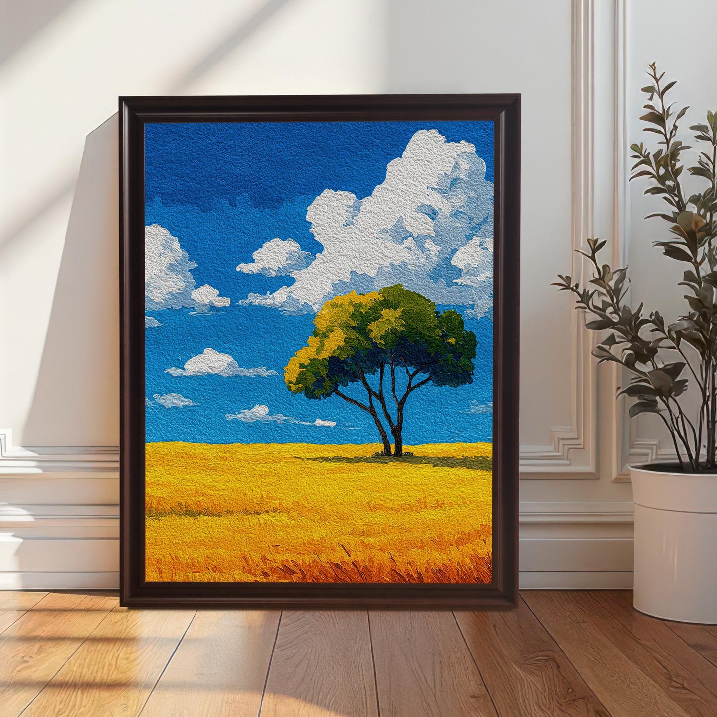 A painting of a tree in a field in a coffee brown frame leaning against a white paneled wall. A potted plant stands to the right of the painting.