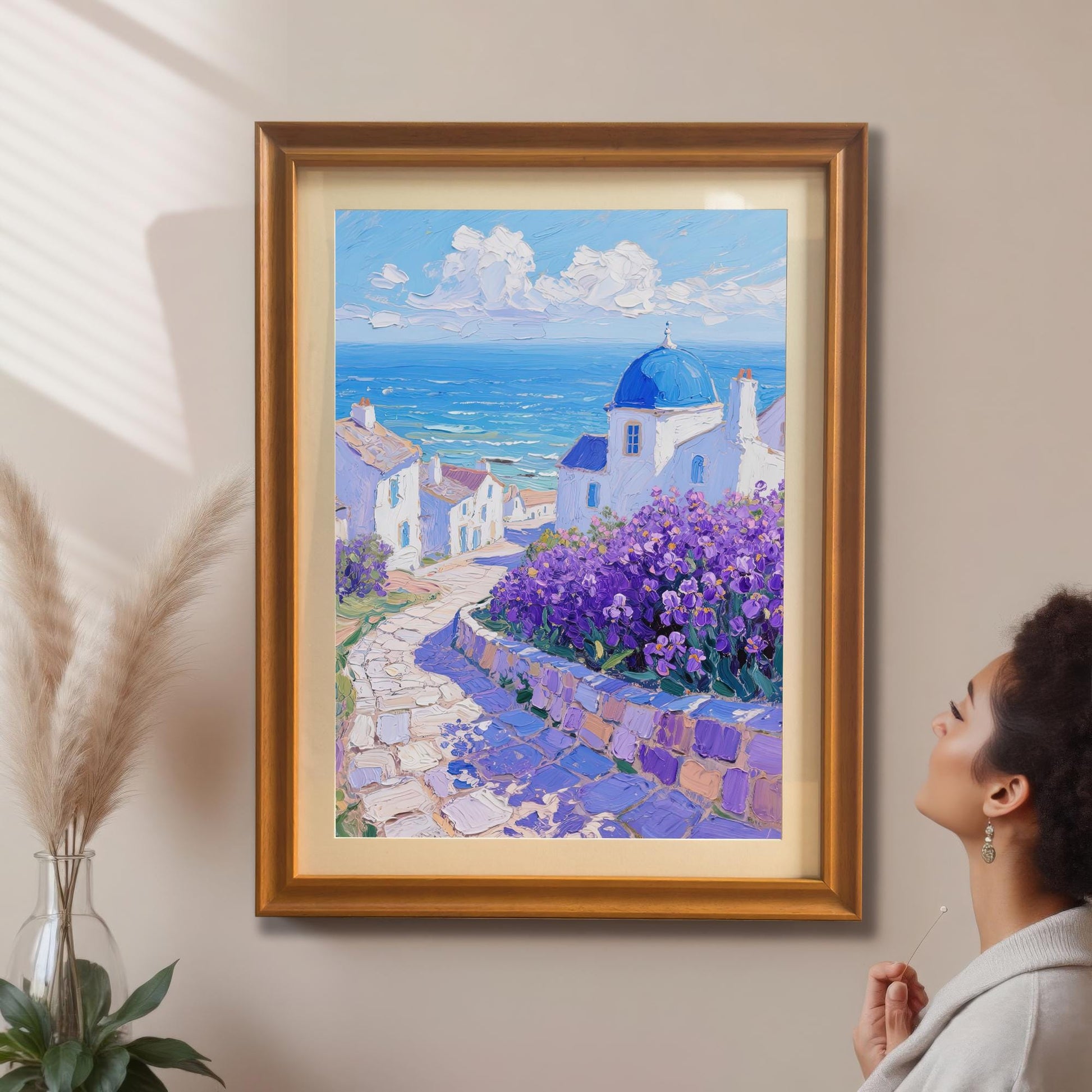A woman looks up at a framed painting of a Greek village with a blue-domed church, mounted on a tan wall. The frame is a warm, orange-gold color with a mount.