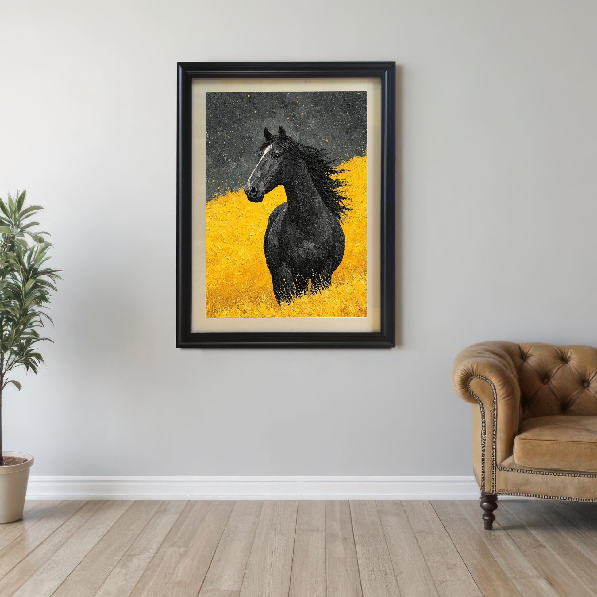 A black-framed painting of a black horse standing in a bright yellow field against a dark grey sky, hanging on a white wall above a leather chair and next to a houseplant.