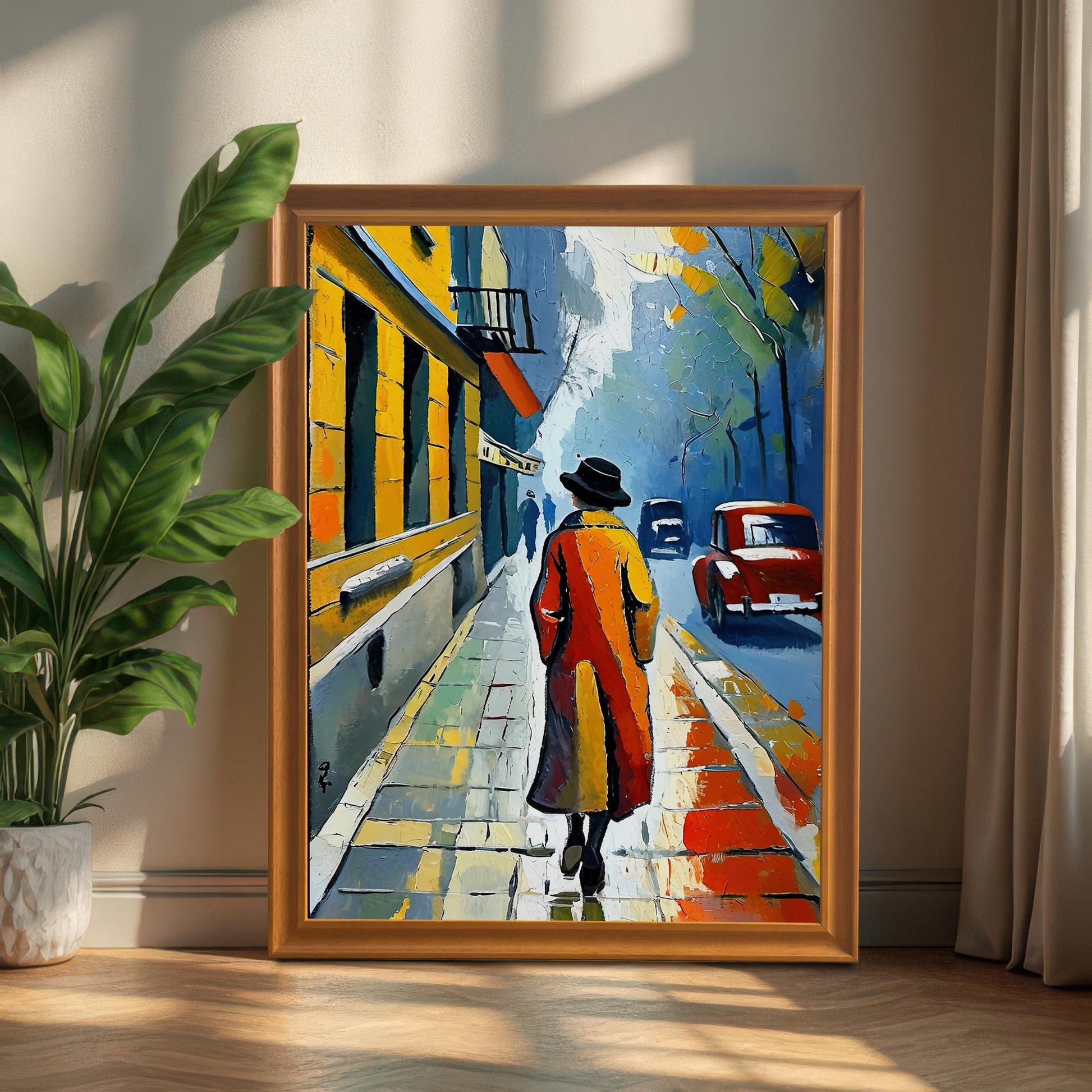 A framed print of a person in a red coat walking down a street. The frame is a light orange wood color and the print is propped on a wooden floor against a wall next to a large potted plant.