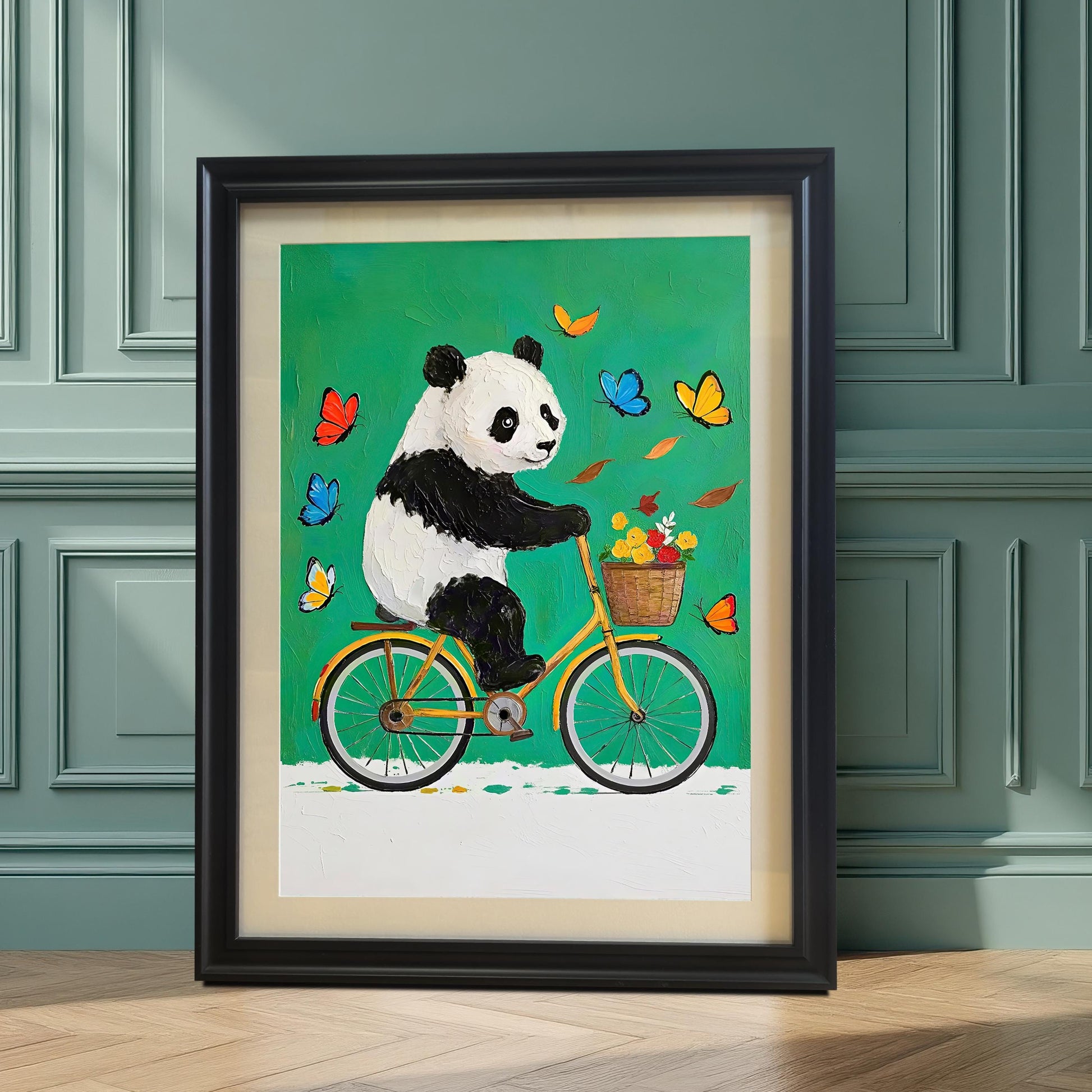A framed art print of a cute panda riding a bicycle is displayed on the floor against a sage green wall with wainscoting. The frame is black with a light-colored mount.