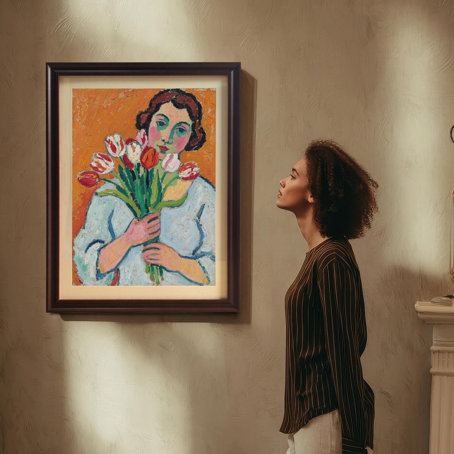 A woman with curly hair looks up at a framed art print hanging on a wall. The print is of a woman holding tulips.
