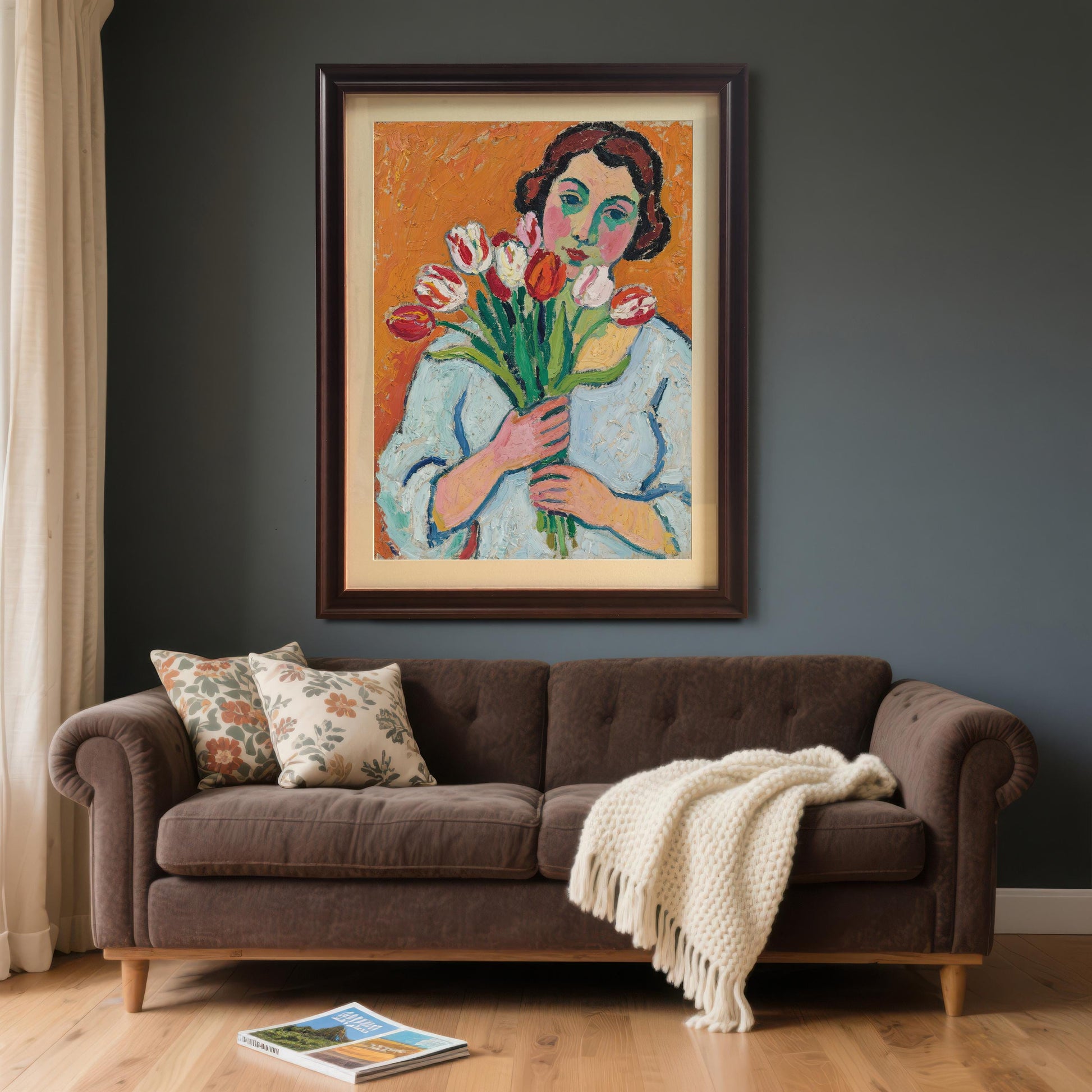 A framed art print of a woman with tulips hanging on a dark gray wall above a brown sofa with two pillows and a white blanket draped over it.