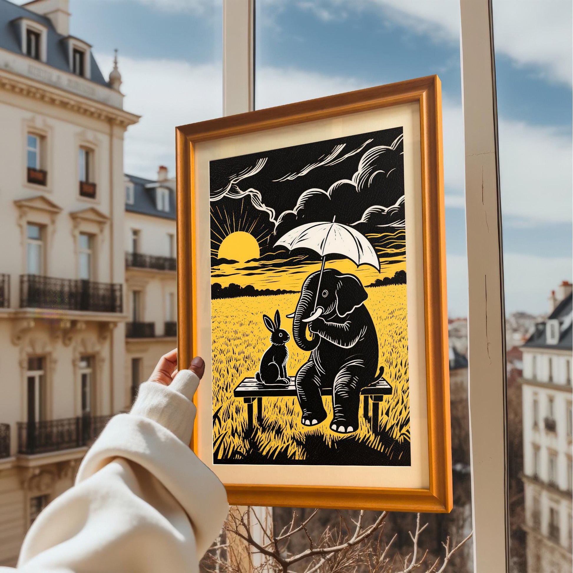 A hand holding an orange-framed print of an elephant and a rabbit sitting on a bench against a bright window view of city buildings.