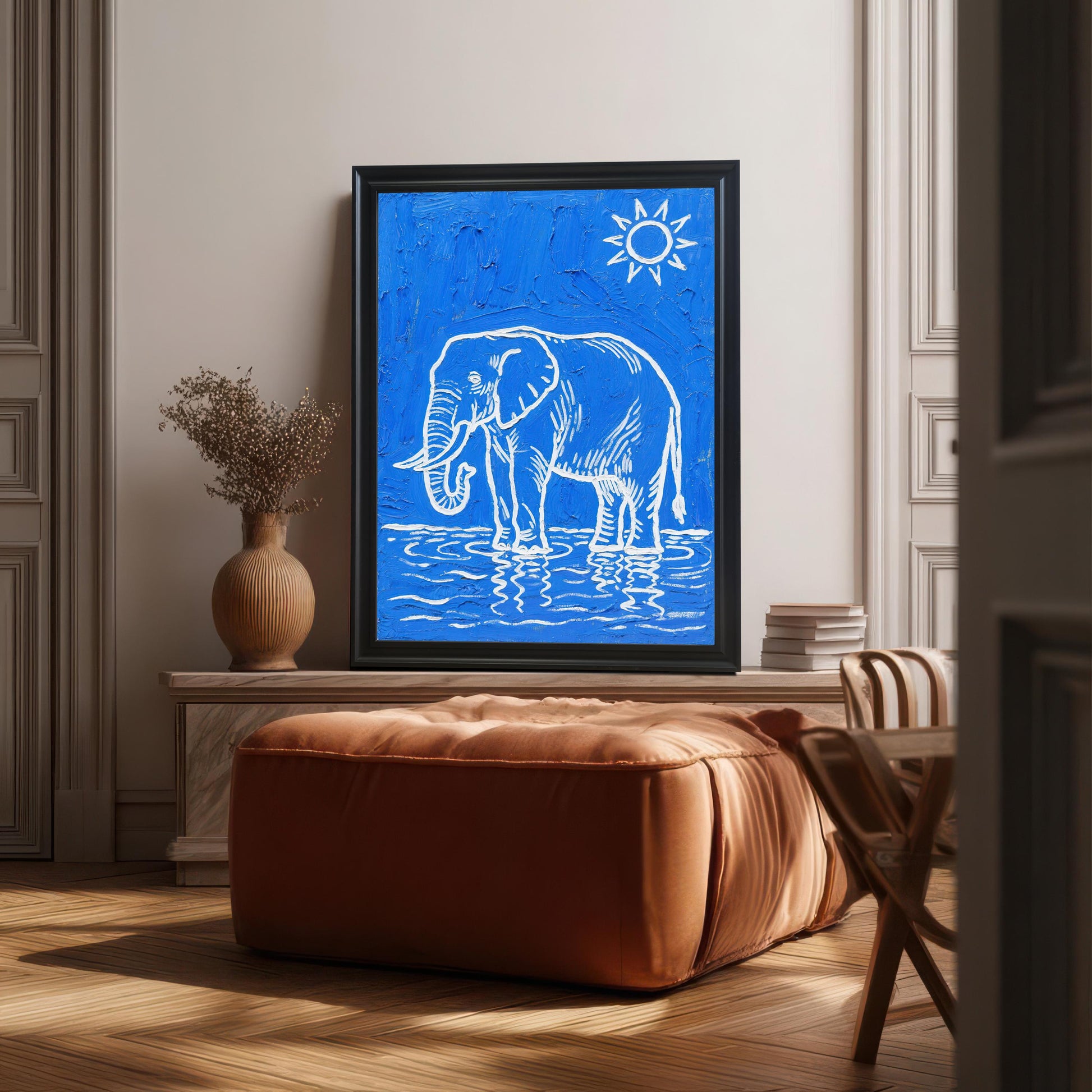 A large framed black and blue elephant print is propped on a low console behind a leather ottoman in a well-lit living space.