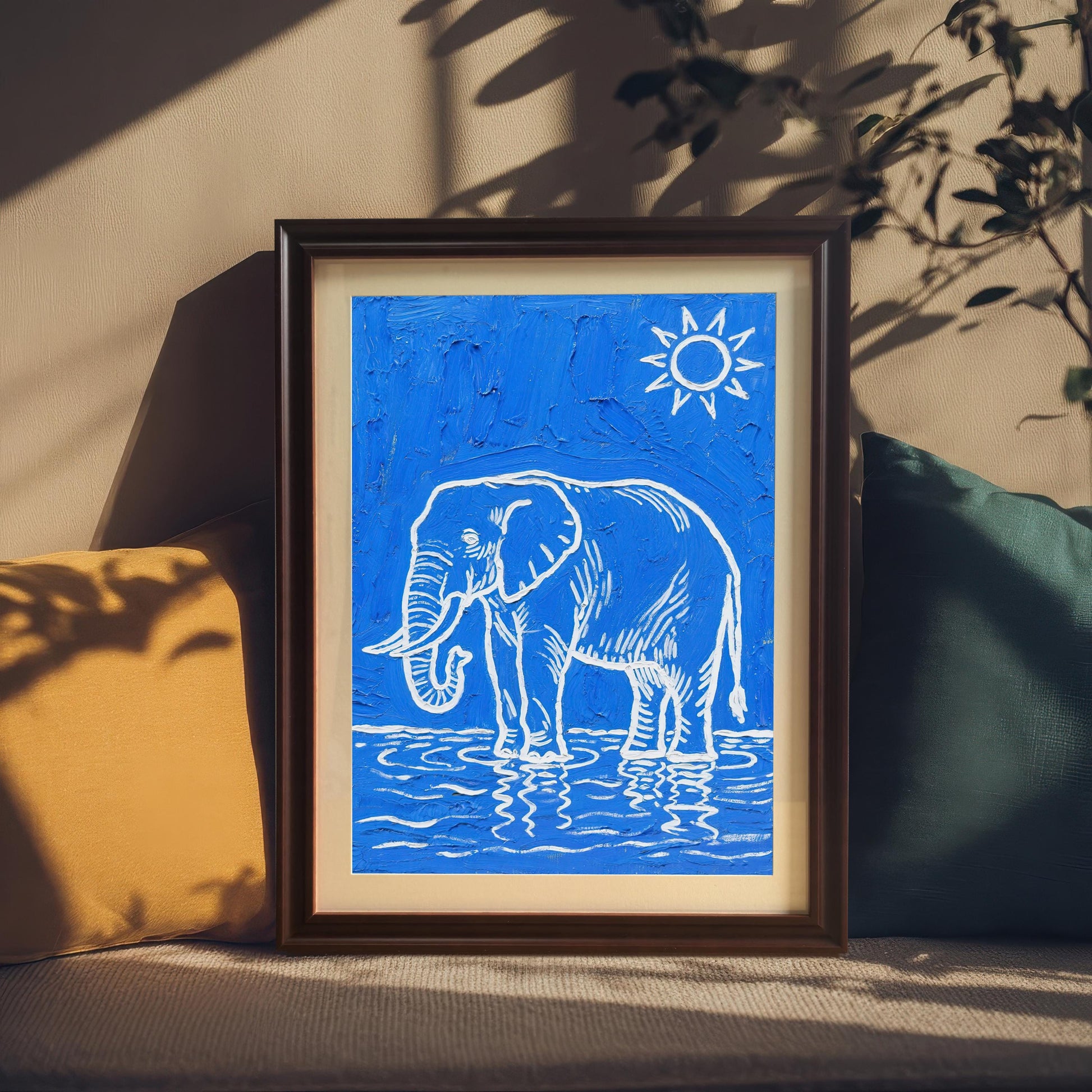 A framed blue and white elephant print stands between a yellow and a teal pillow in a sunlit room.