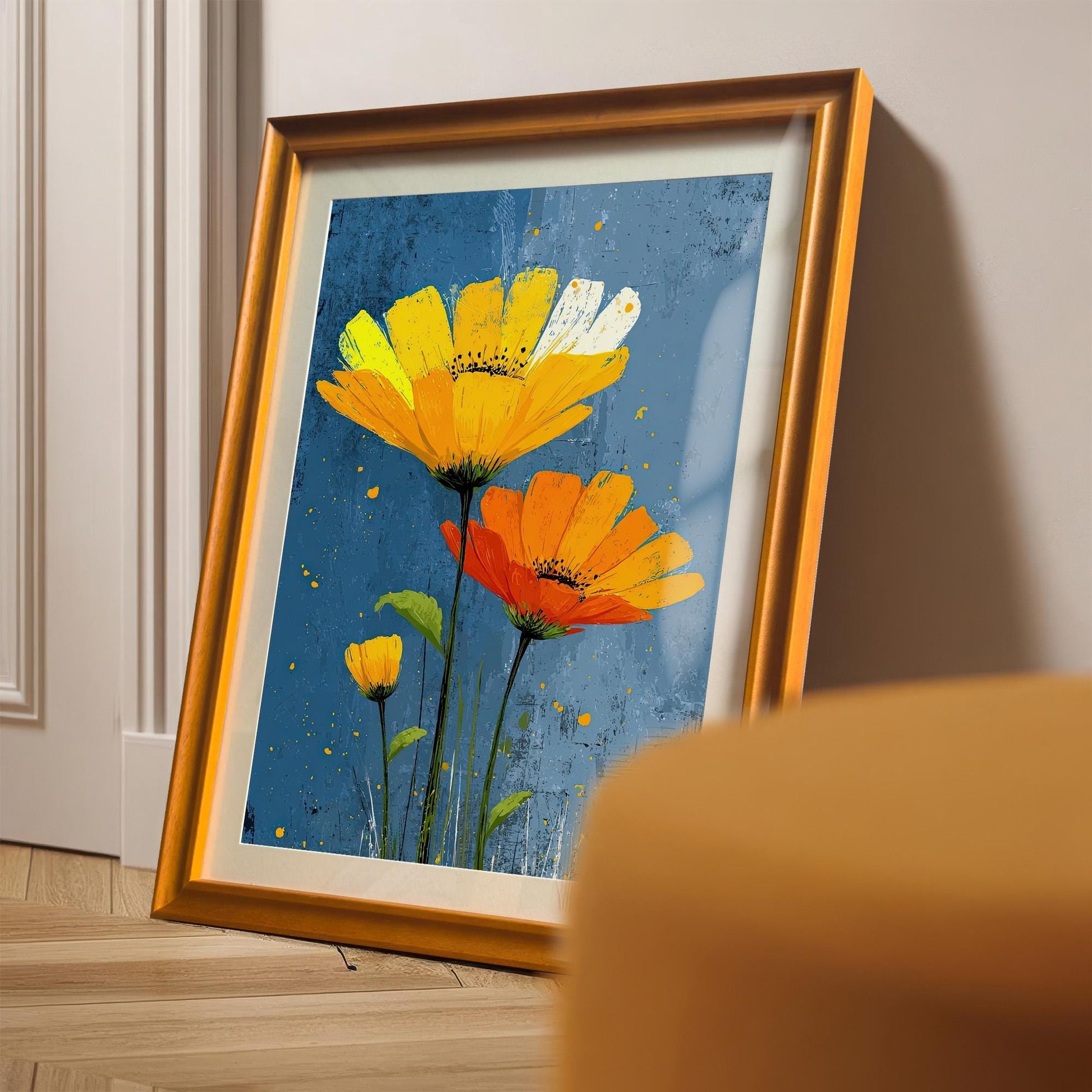 A framed painting of yellow and orange flowers on a blue background is leaning against a white wall on a wooden floor, with an orange chair in the foreground.