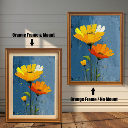Two framed art prints of yellow and orange flowers on a wooden floor. The print on the left is in an orange frame with a mount, and the one on the right is in an orange frame without a mount.