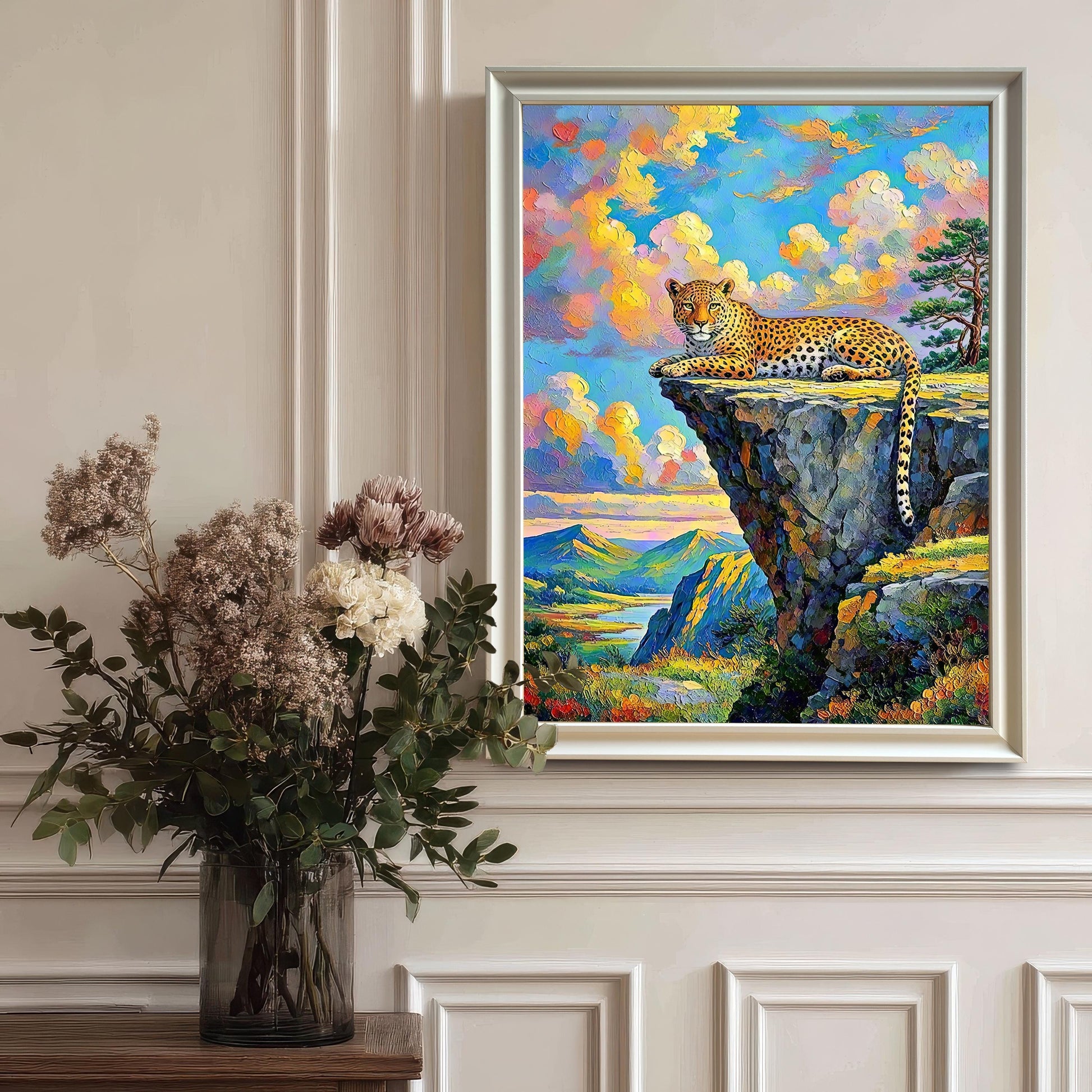 Framed wall art of a leopard on a cliffside, hanging on a white paneled wall with a vase of flowers on a table below it.