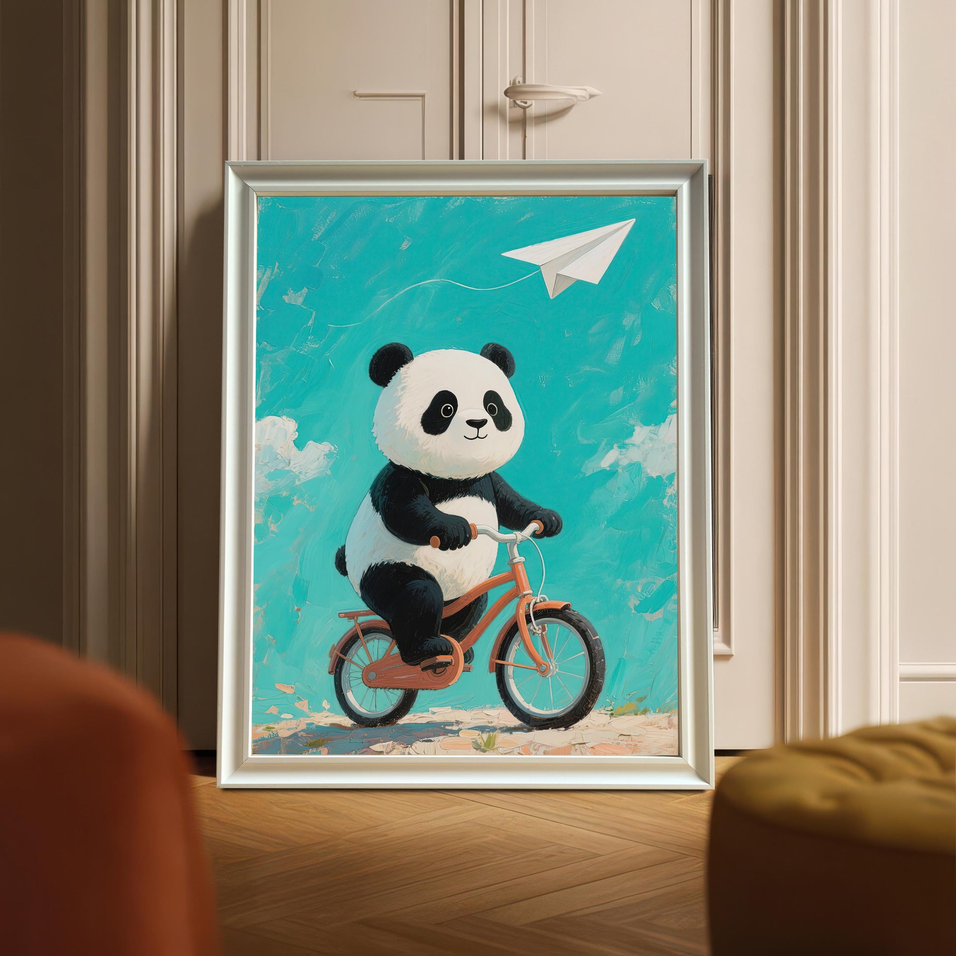 A framed print of a panda on a bicycle leaning against a white wardrobe. The frame is a silvery-white color. The image is a full shot, showing the framed print in an indoor setting.
