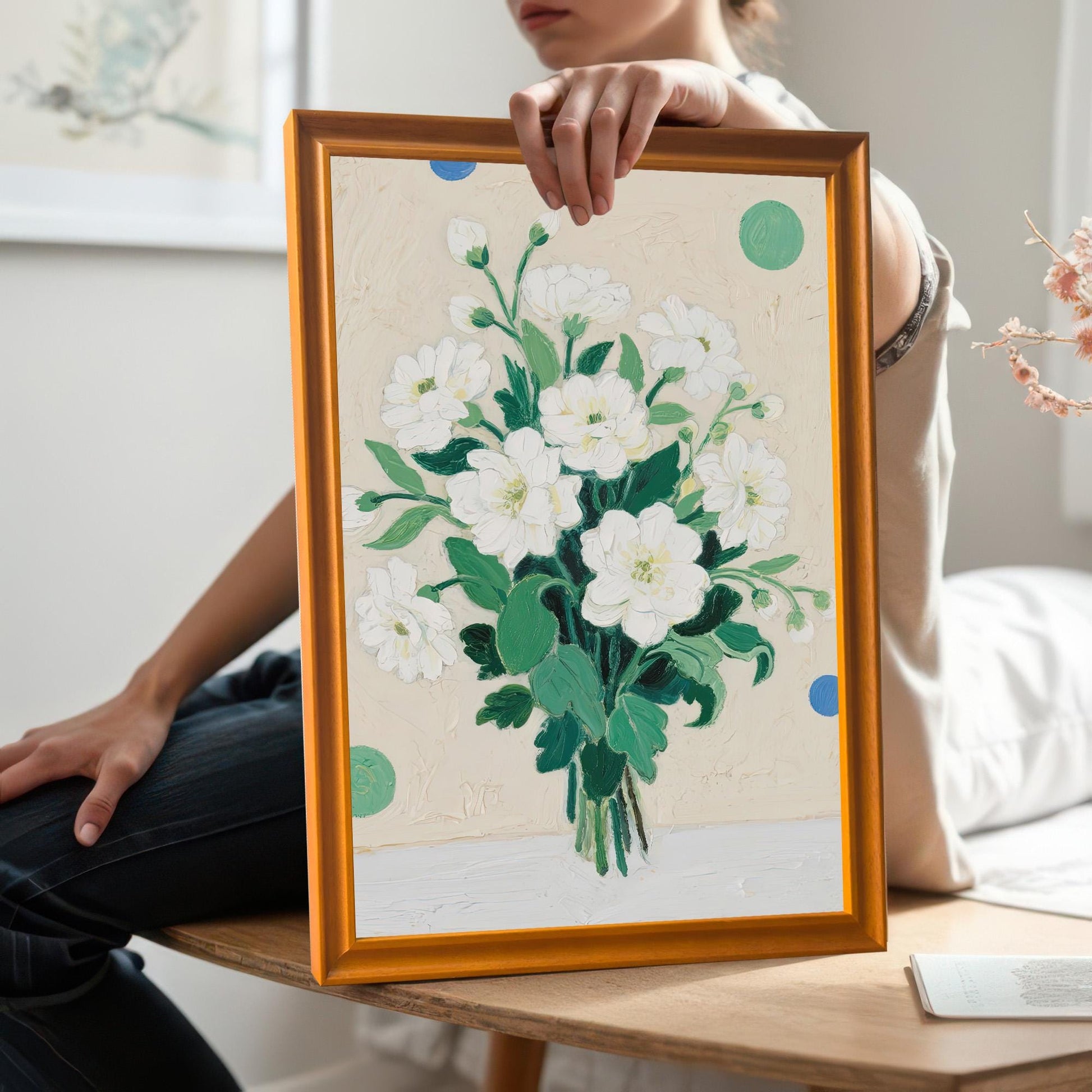 A person holds a framed painting of a bouquet of white flowers. The frame is a light, natural wood color. The person is sitting on a wooden bench in a sunlit room.