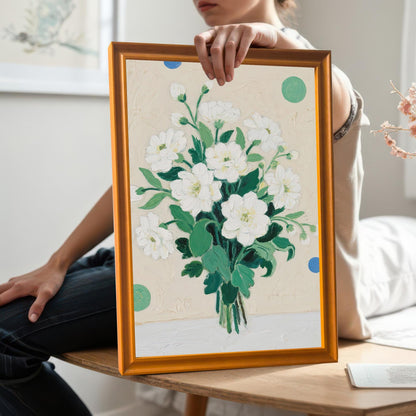 A person holds a framed painting of a bouquet of white flowers. The frame is a light, natural wood color. The person is sitting on a wooden bench in a sunlit room.