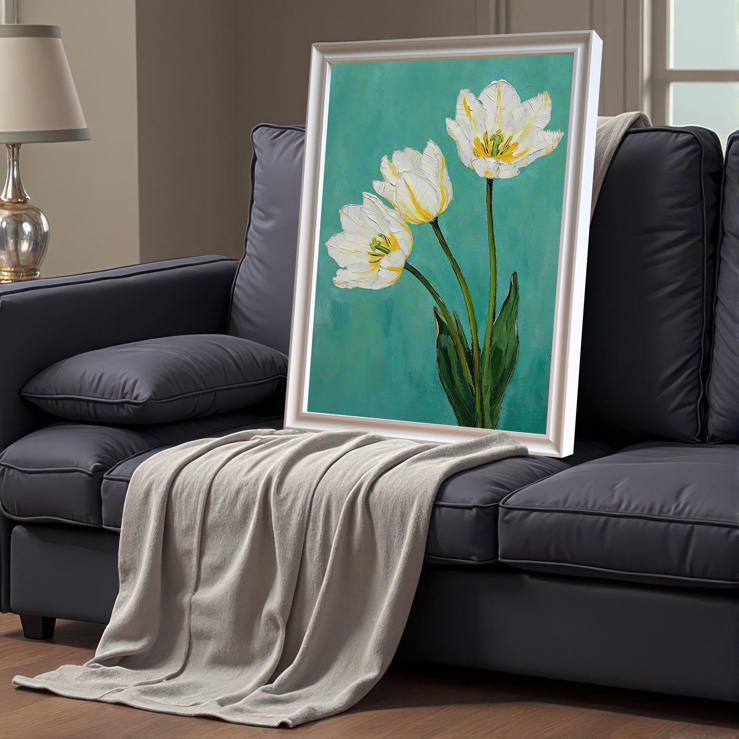 A framed art print of three white tulips with green leaves on a teal background, leaning on a dark gray couch. The print has a white frame and a white mat. A gray blanket is draped on the couch.