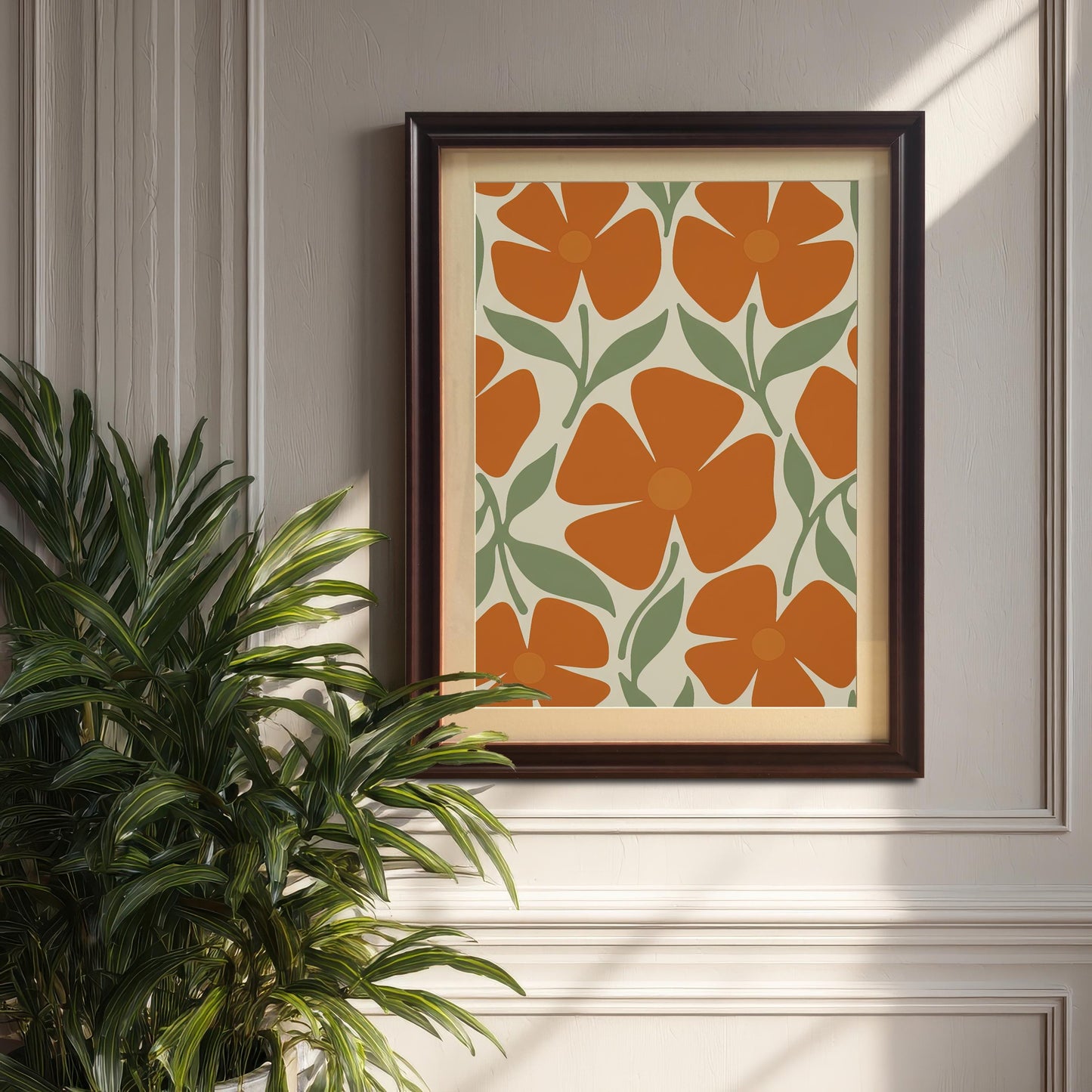 A framed art print with a repeating pattern of orange flowers and green leaves hanging on a white wainscoting wall, with a large potted plant in the foreground.