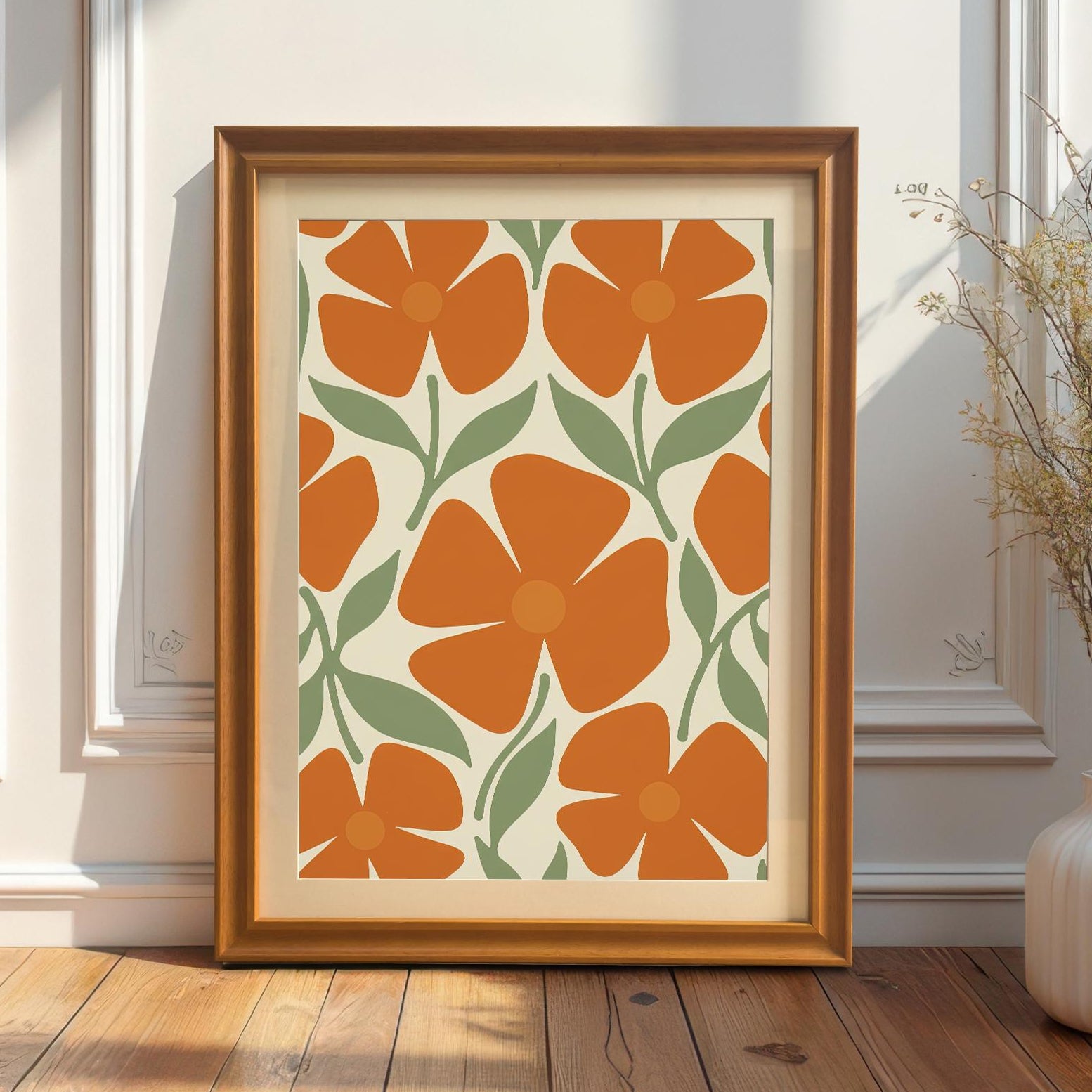 A framed art print with a repeating pattern of orange flowers and green leaves, with a gold frame, leaning on a wooden floor against a white wainscoting wall.