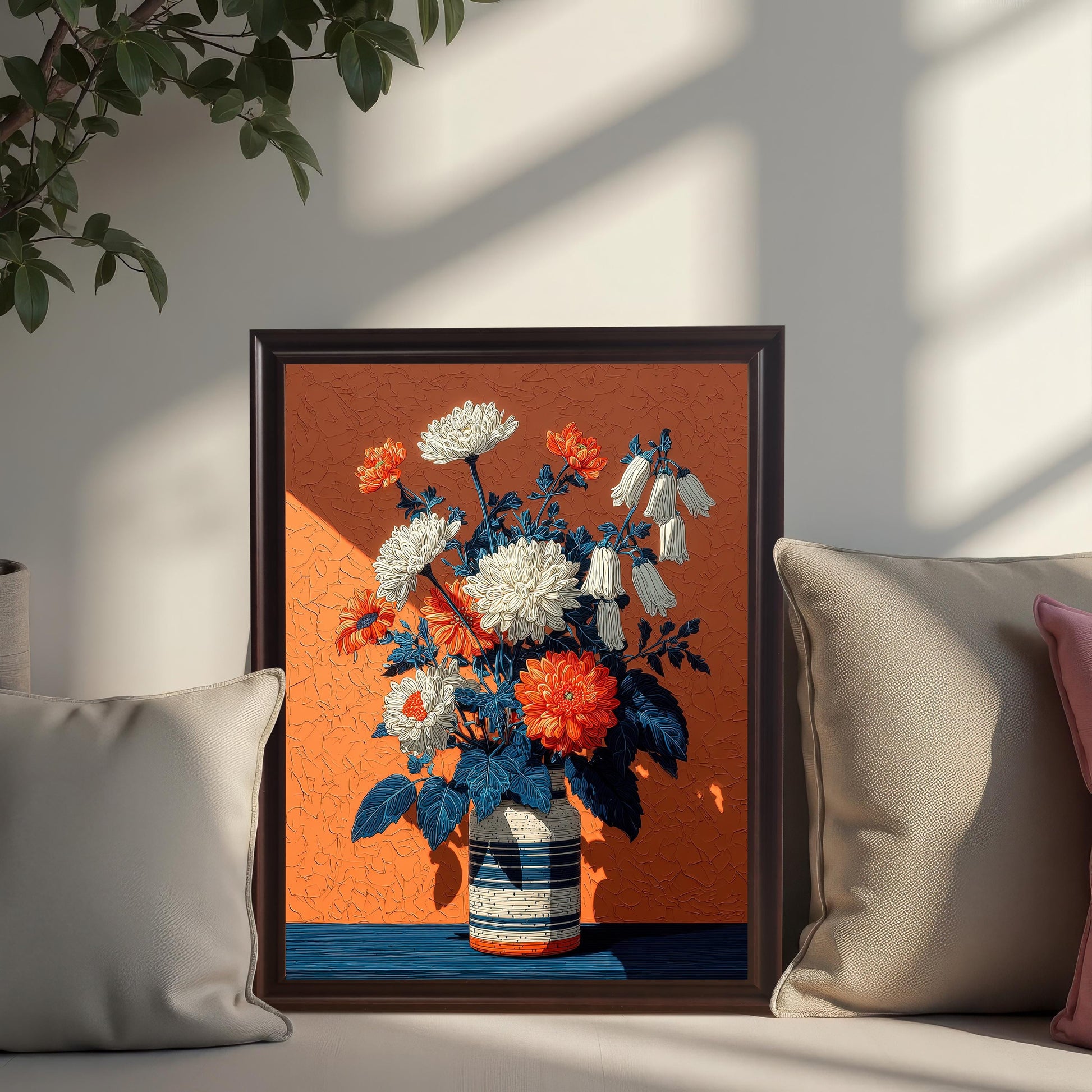 A framed, dark-framed art print of a floral arrangement in a vase on an orange background, propped between two pillows on a white couch.