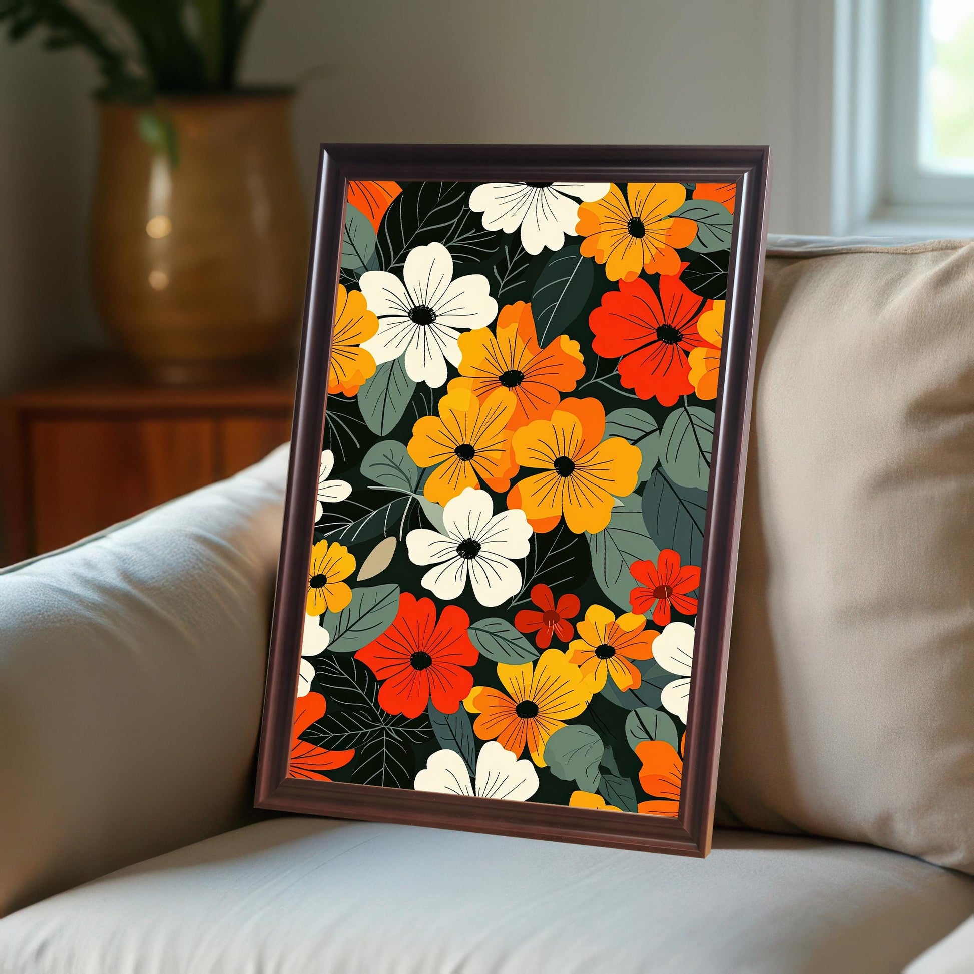 A coffee-colored, framed art print with colorful flowers leans on a beige couch with a large plant pot in the background.