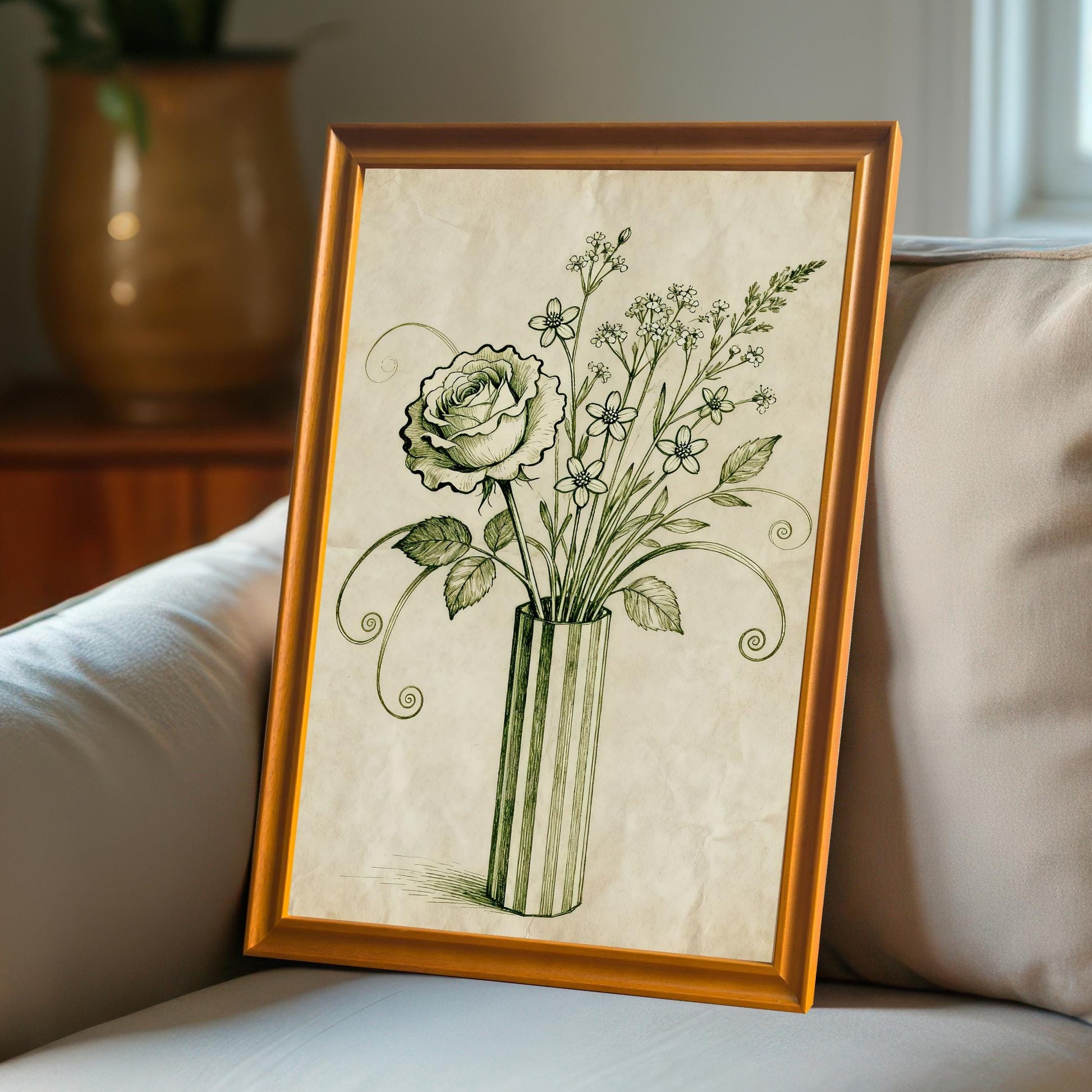 A framed art print of a vintage-style rose and flowers in a vase leans against a couch with a beige pillow. A window and large plant pot are blurred in the background.