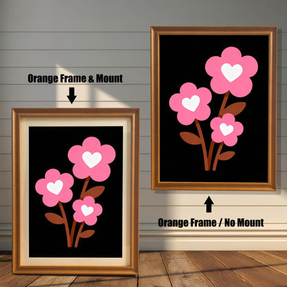 A framed art print of three pink flowers with white hearts is shown in two styles against a wooden wall. One has an orange-colored frame with a white mount, the other is a larger size with the same frame, but no mount.