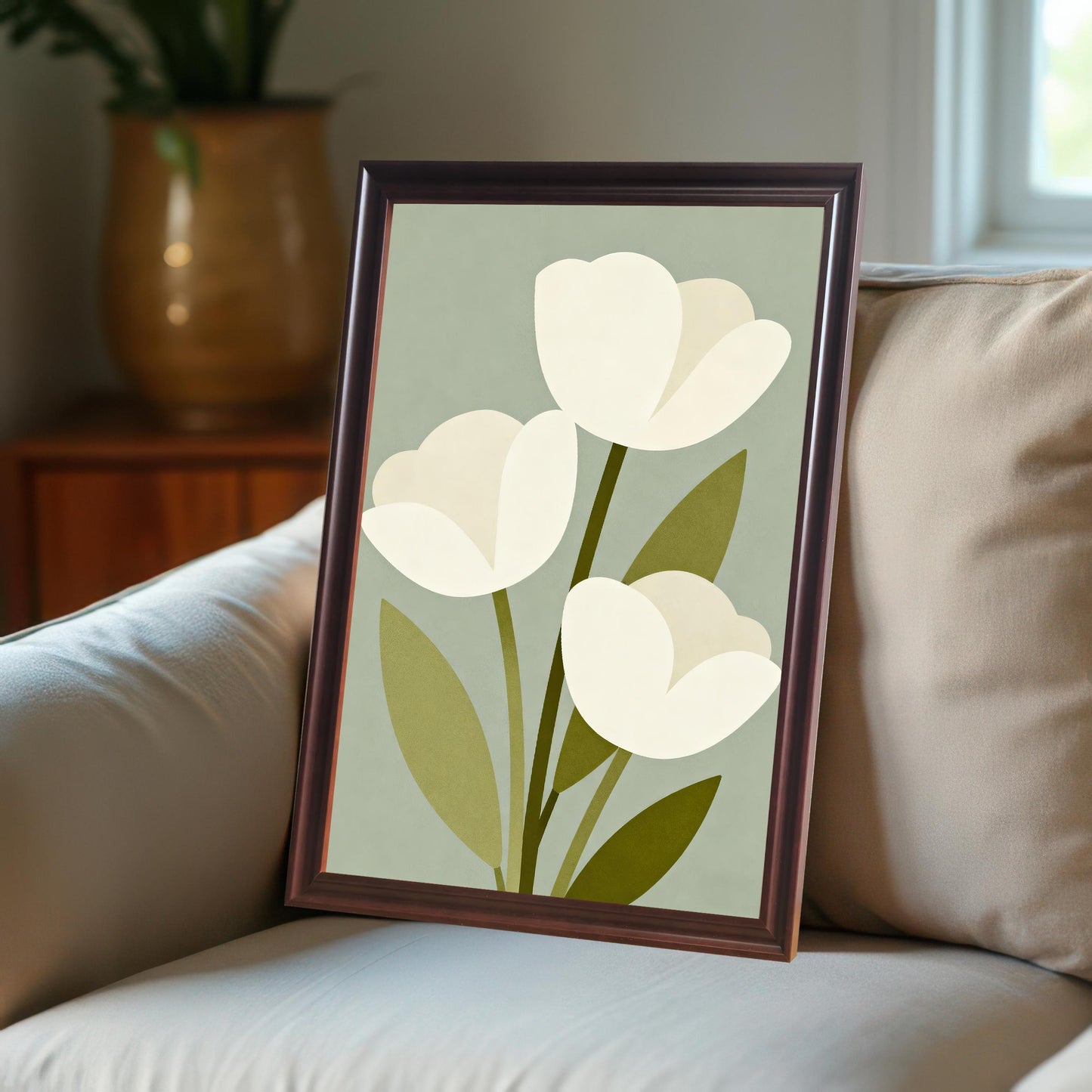 A framed art print of three white tulips leans against a couch with a beige pillow. A window and large plant pot are blurred in the background.