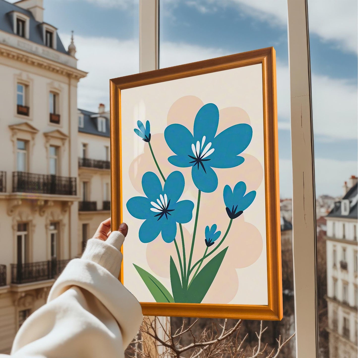 A hand holds a framed painting of stylized blue flowers in front of a window overlooking a cityscape with apartment buildings and a cloudy sky.