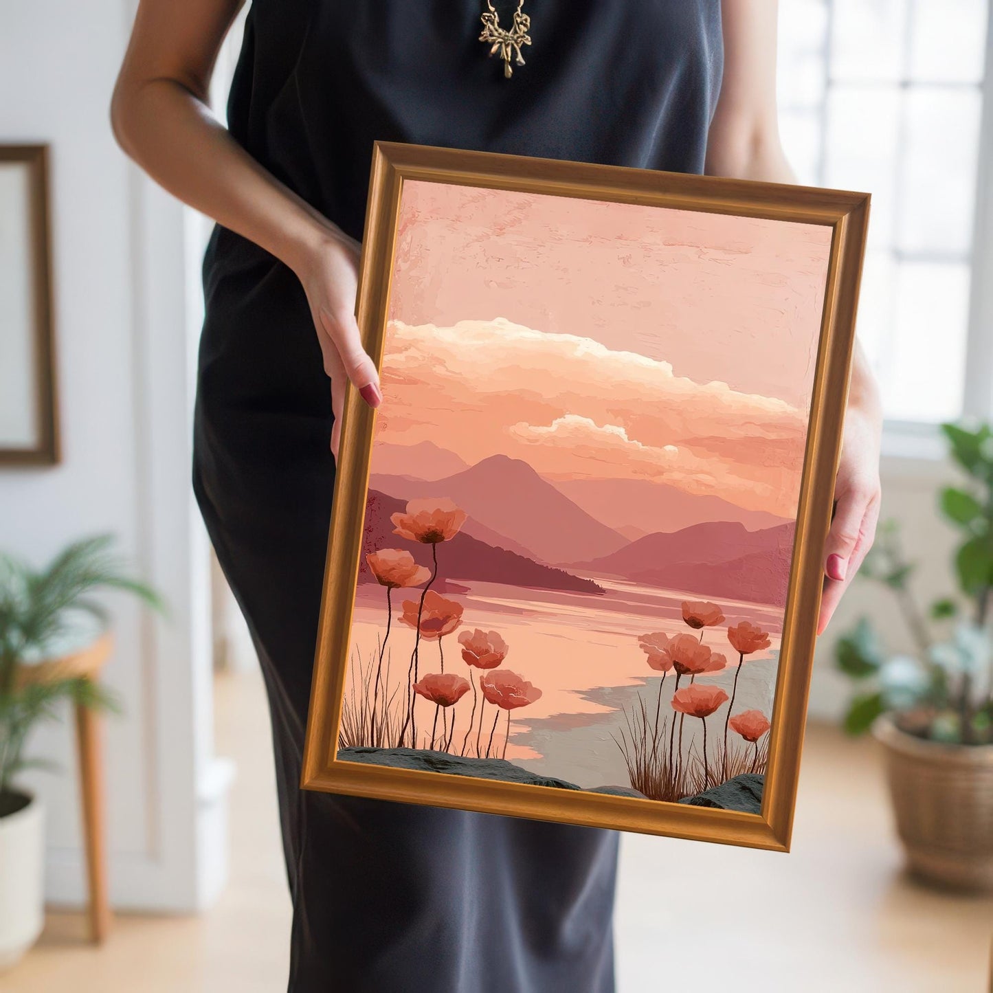 A person in a black dress holding a framed print of the poppy landscape art. The frame is a shiny gold or brass color, and the art depicts a pink and orange sunset over mountains and a lake with poppies in the foreground.