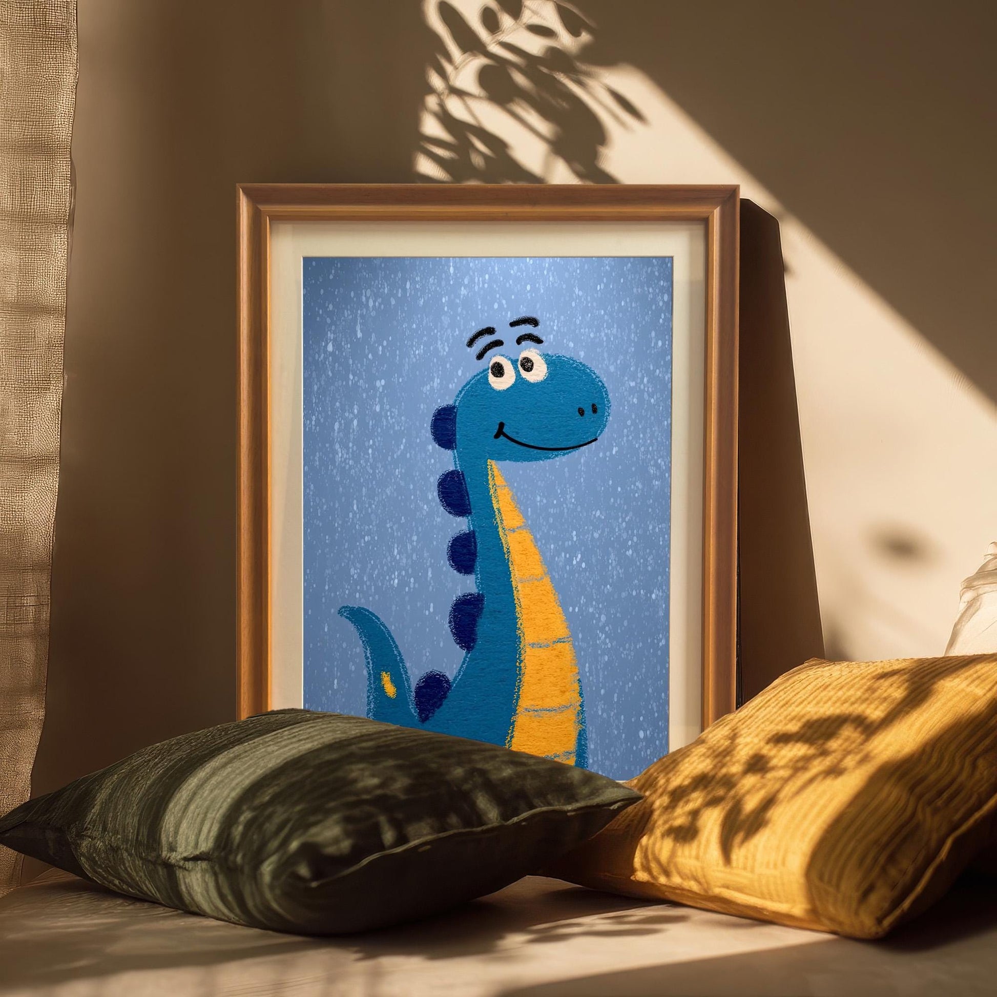 A cozy close-up of the framed children's art print of a blue and orange cartoon dinosaur, framed in light brown wood, leaning against a wall or piece of furniture. It is surrounded by soft throw pillows, one dark green and one yellow. Sunlight streams from the top, casting dramatic shadows of leaves and window frames across the scene.