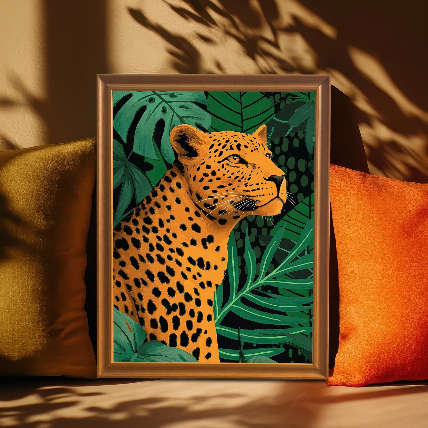 Close-up, cozy lifestyle shot of the framed leopard print leaning against a wall, surrounded by an olive-green pillow and an orange pillow. The print is in a light brown/orange wood frame. Strong sunlight casts shadows of leaves across the wall and art.