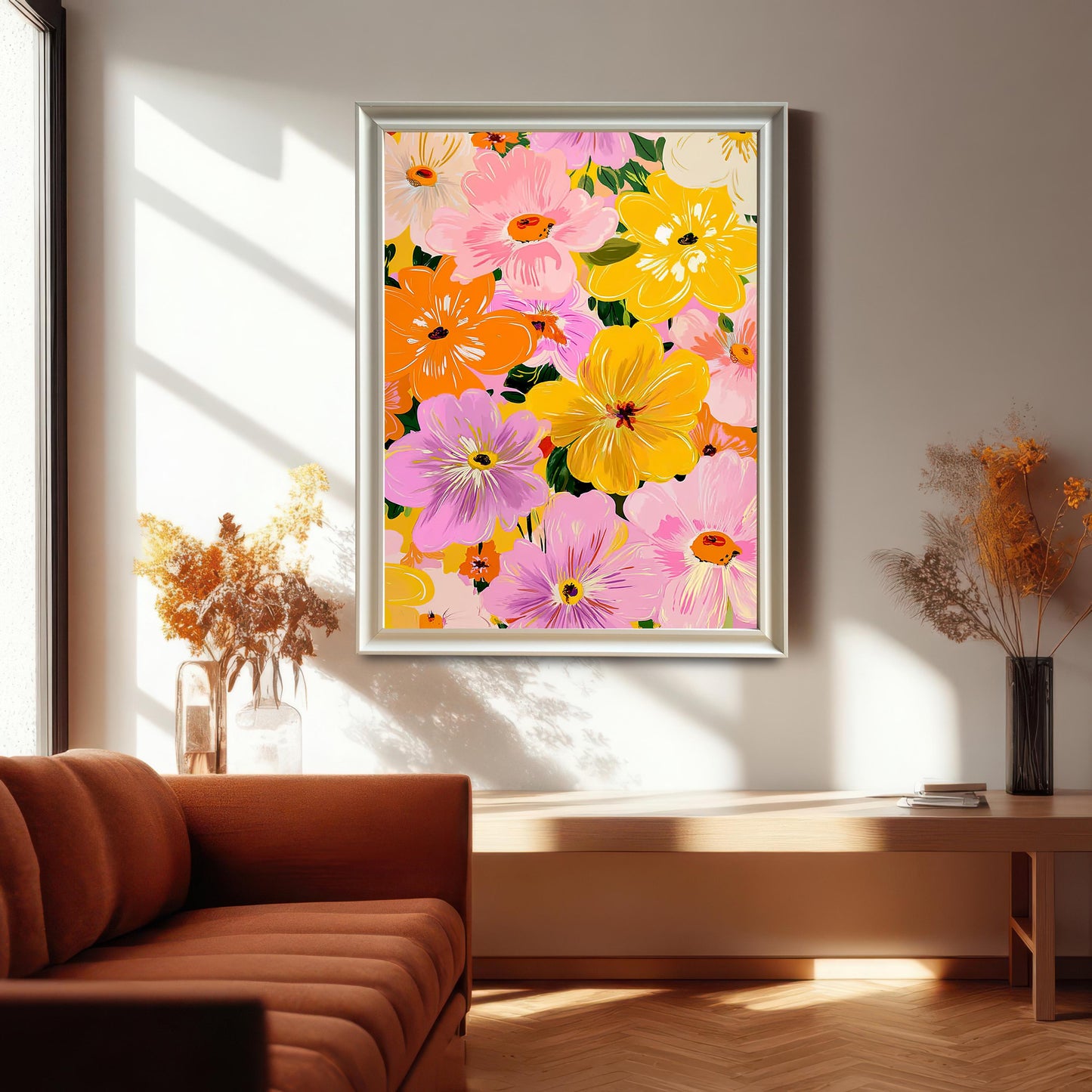Lifestyle photo of the framed art print featuring a dense, colorful pattern of flowers in pink, yellow, orange, and purple. The print is hanging on a bright wall above a wooden console and is framed in a light-colored/silver frame. A rust-orange sofa is visible on the left.