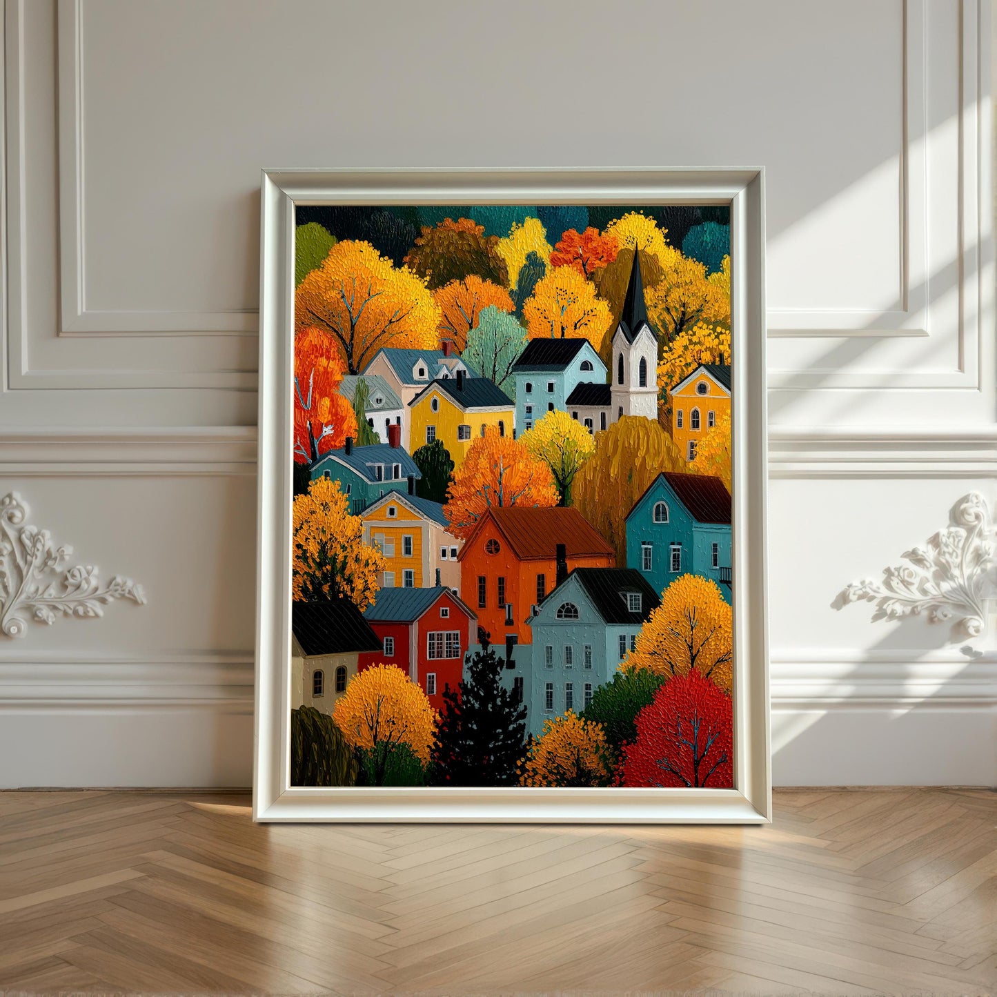 A framed print of the autumn village scene in a white frame, leaning against a white decorative wainscoting wall and wood floor.