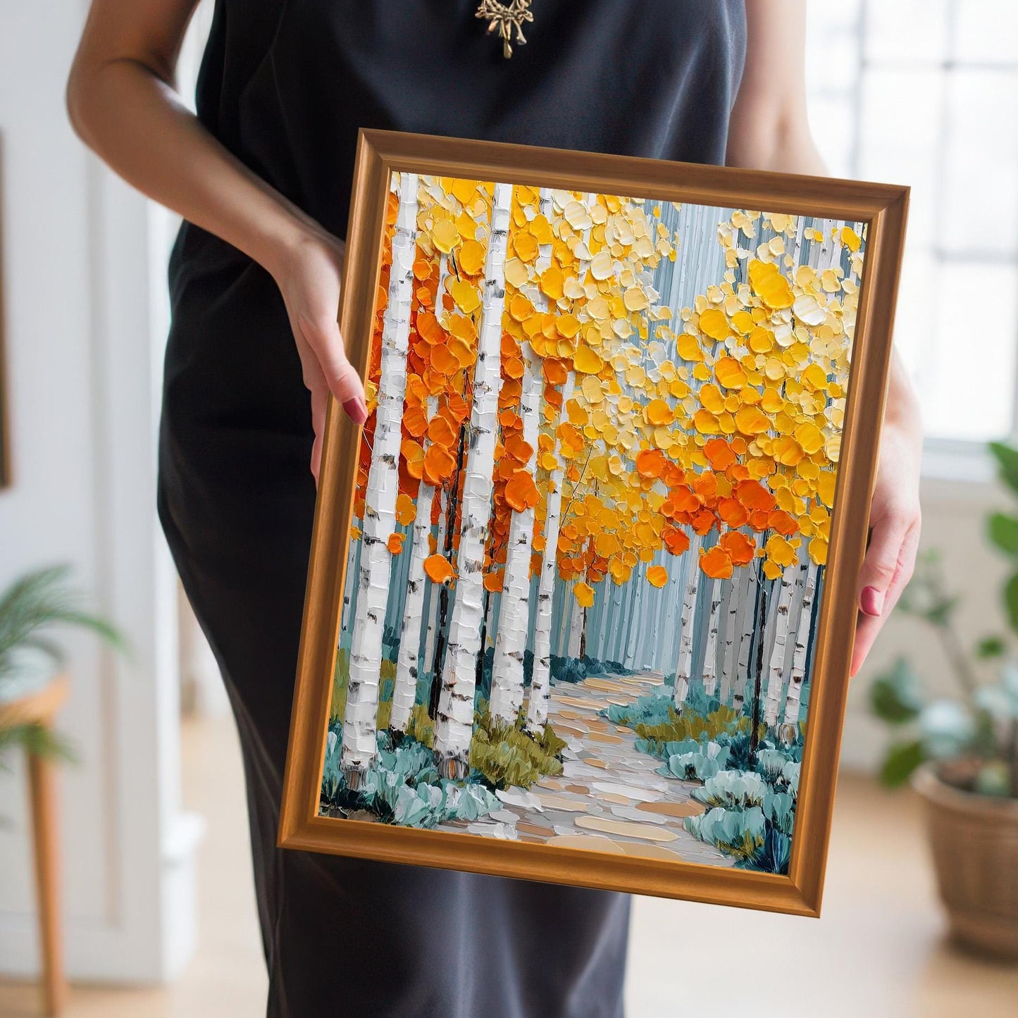 A close-up of a person in a black dress holding a framed print of an autumnal aspen forest. The frame is a gold/bronze color. The art features a path winding through white birch trees with textured orange and yellow foliage.