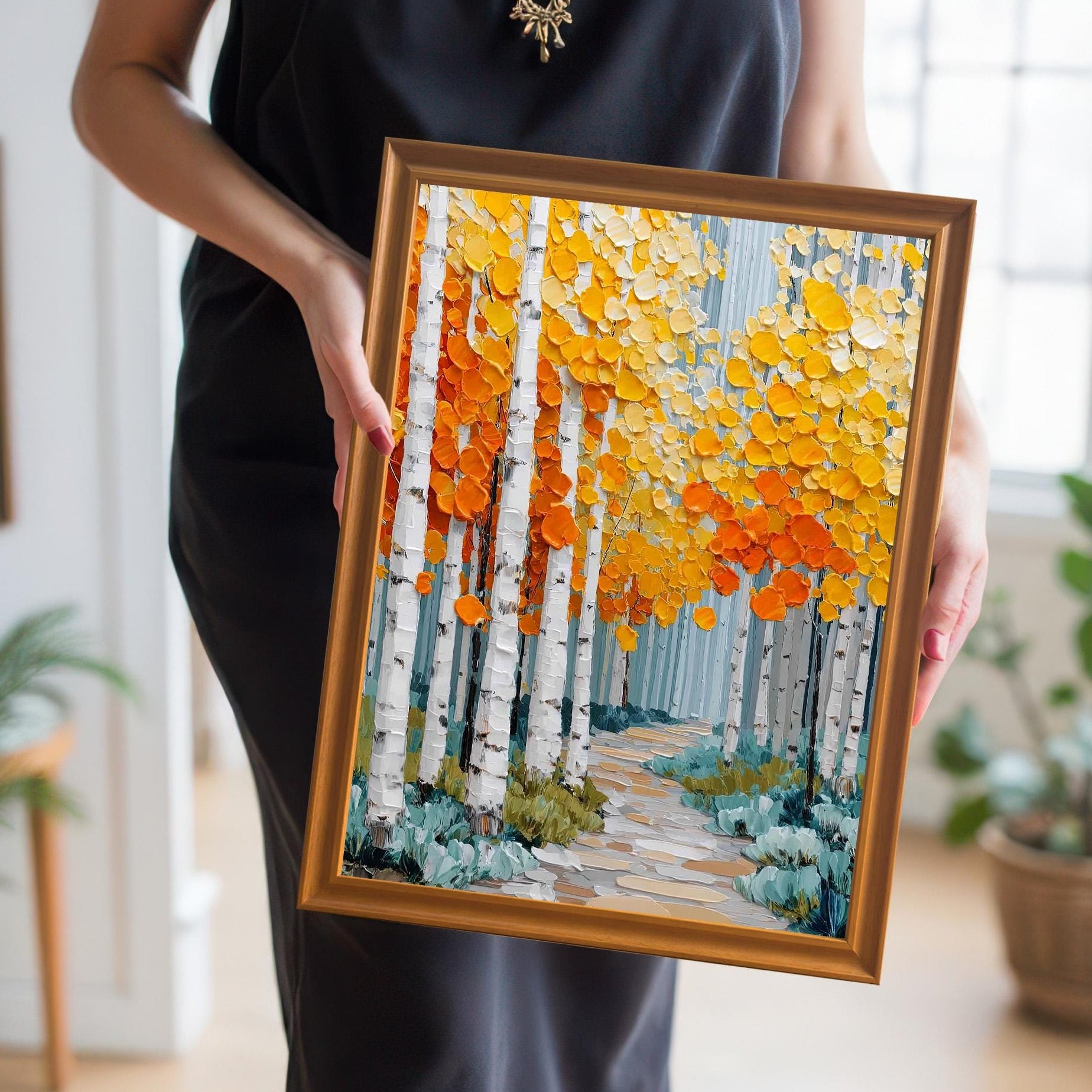 A close-up of a person in a black dress holding a framed print of an autumnal aspen forest. The frame is a gold/bronze color. The art features a path winding through white birch trees with textured orange and yellow foliage.