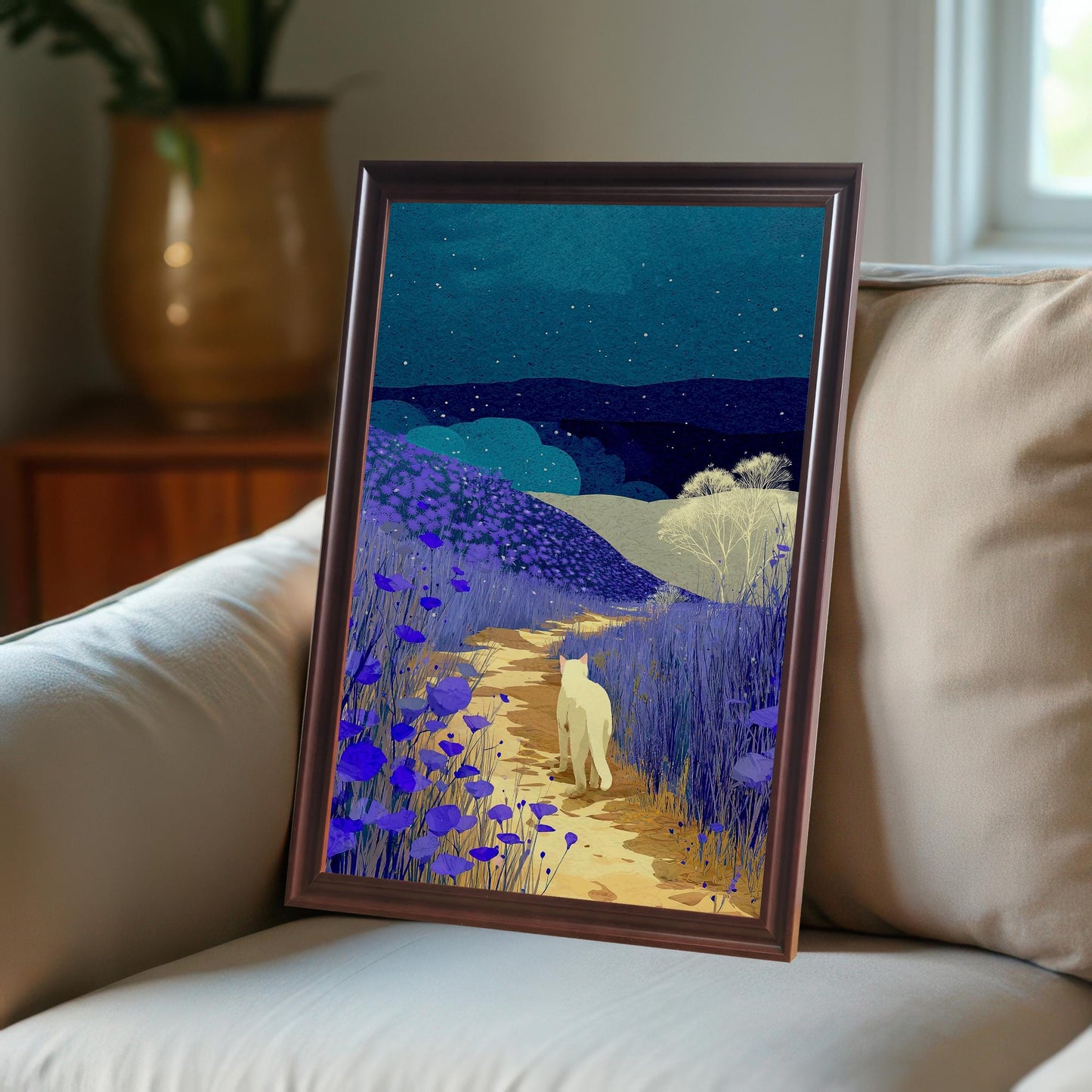 Framed print of a white cat walking on a path through fields of purple flowers under a starry, dark blue night sky. The print is leaning against a light-colored couch. The frame is dark brown or coffee-colored.