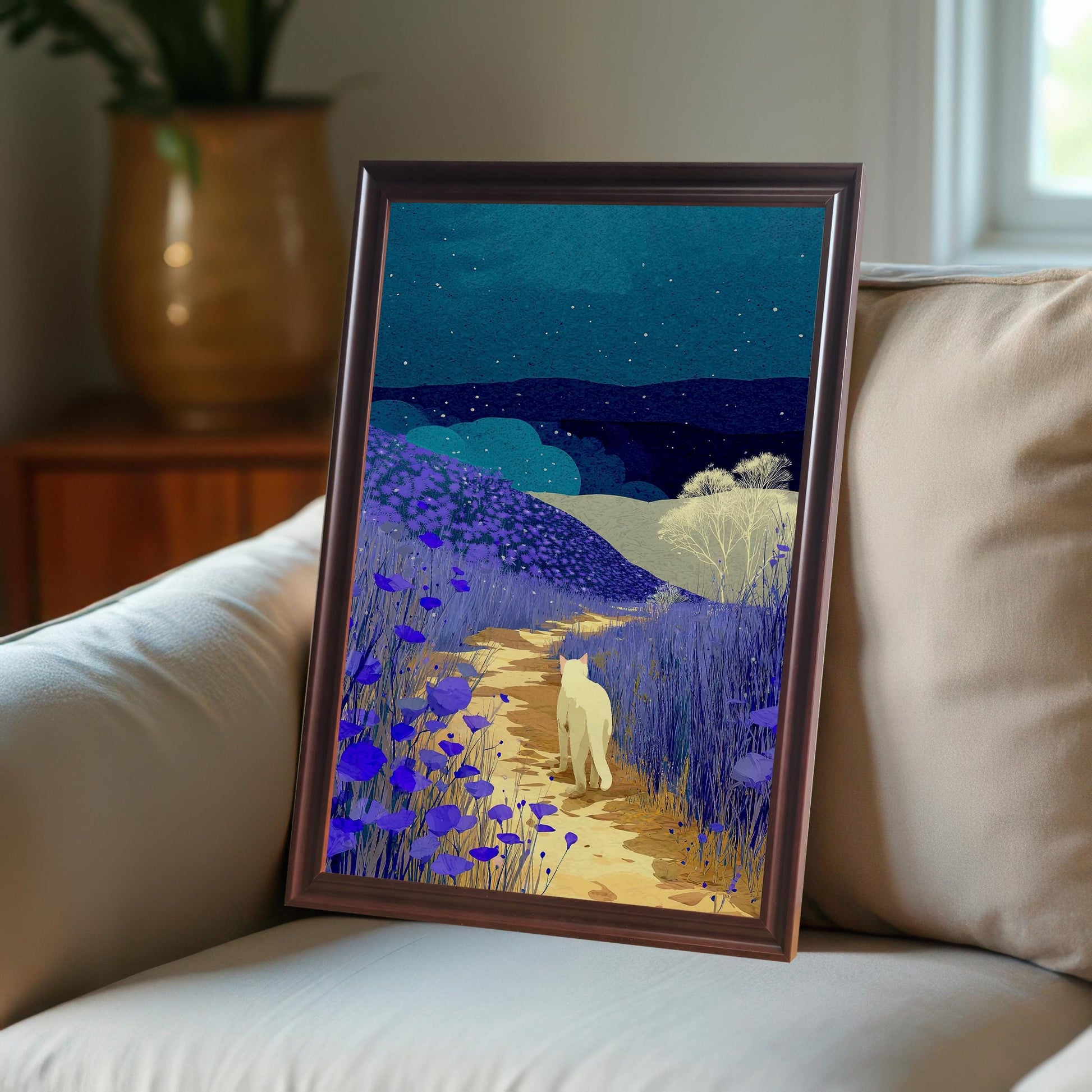 Framed print of a white cat walking on a path through fields of purple flowers under a starry, dark blue night sky. The print is leaning against a light-colored couch. The frame is dark brown or coffee-colored.