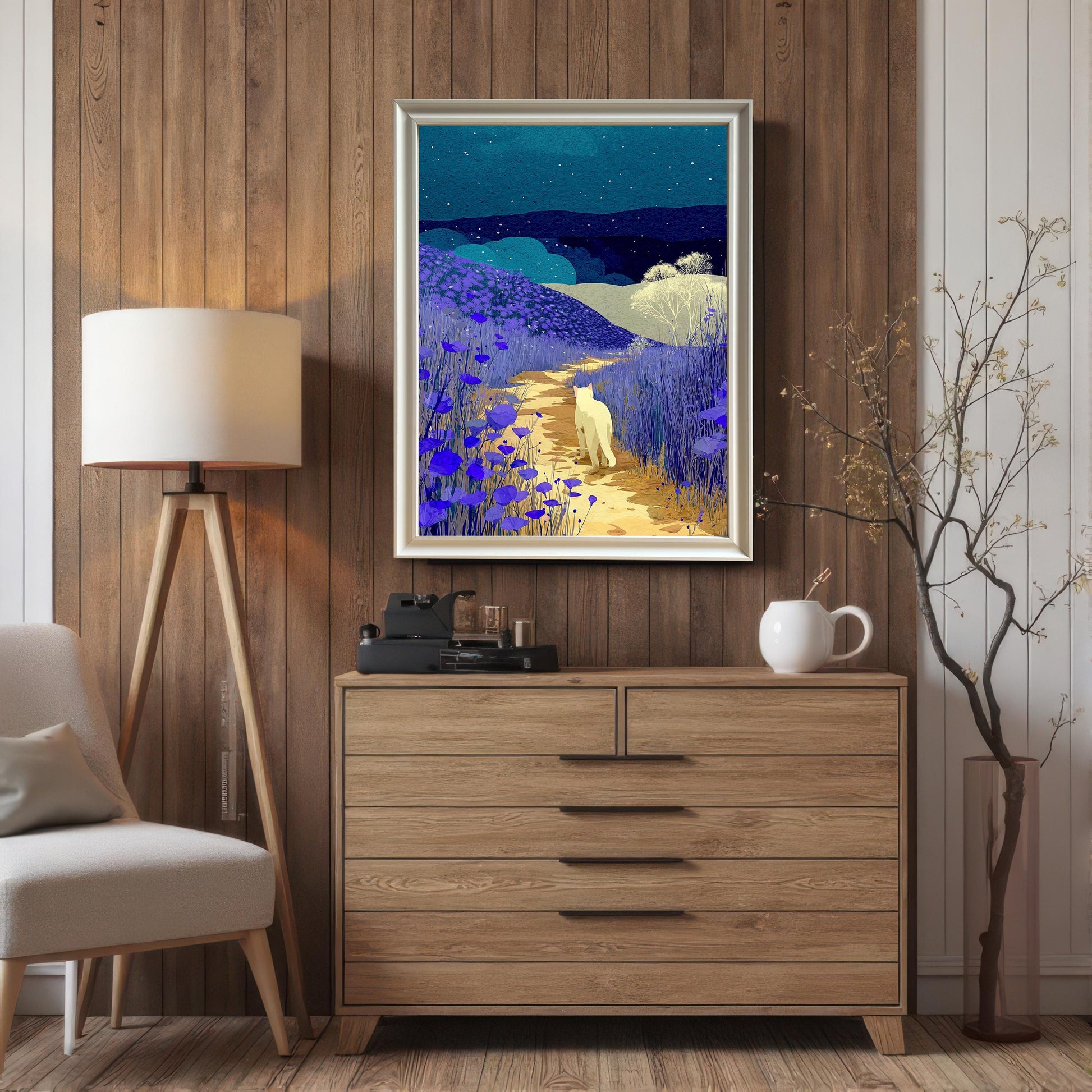 Framed print of the night landscape with the white cat hanging on a vertical wood-paneled wall, centered above a light wood dresser. The frame is a thin silver or light metallic color and includes a white mount (matting). A tripod floor lamp is visible to the left.
