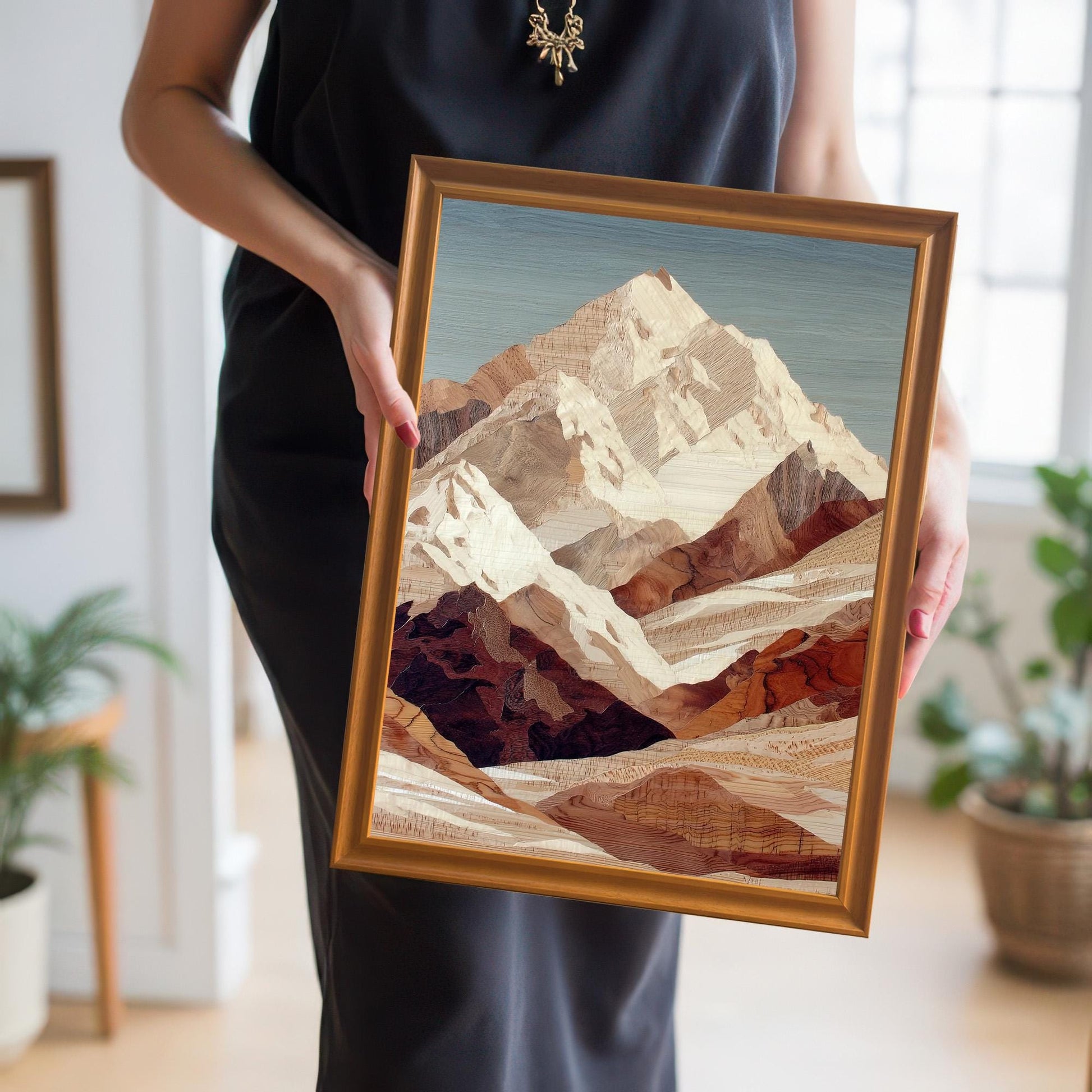 A person in a black dress holds the framed mountain art print at chest level. The frame is a bright orange-gold. The background is a bright, well-lit interior space.