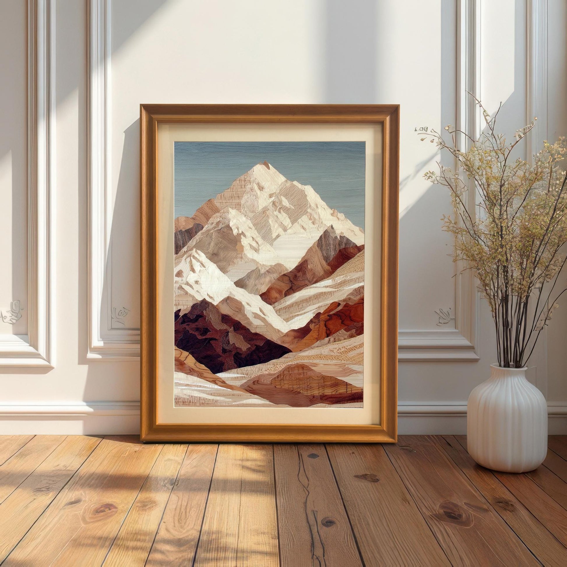 A framed mountain art print is leaning on the floor against a white wainscoted wall. The frame is orange-gold and includes a light-colored mount. A tall white vase with dried branches sits to the right, and sunlight streams in from the left.
