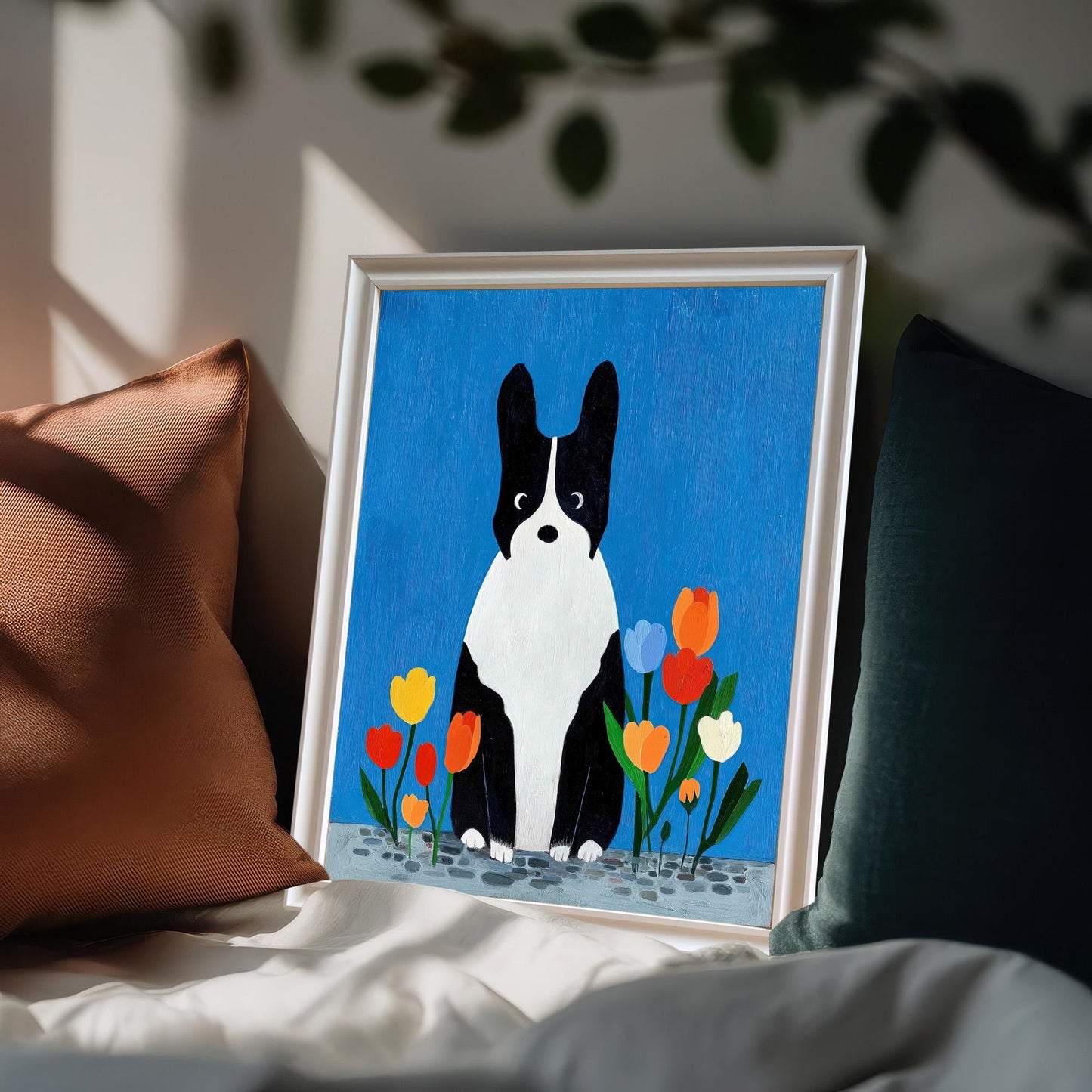 White framed art print of the black and white dog with tulips resting on a bed among dark and brown pillows, suggesting a cozy bedroom setting.