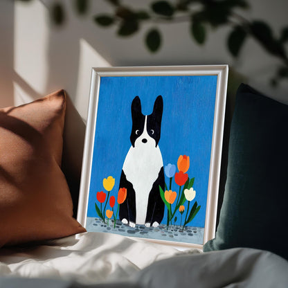 White framed art print of the black and white dog with tulips resting on a bed among dark and brown pillows, suggesting a cozy bedroom setting.