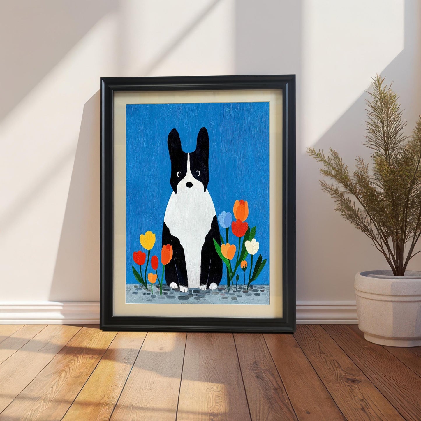 Framed art print of a black and white dog sitting among colorful tulips against a vibrant blue background. The print is in a black frame with a light-colored mat/mount, resting on a wooden floor against a white wall with sunlight streaming across it, next to a potted plant.