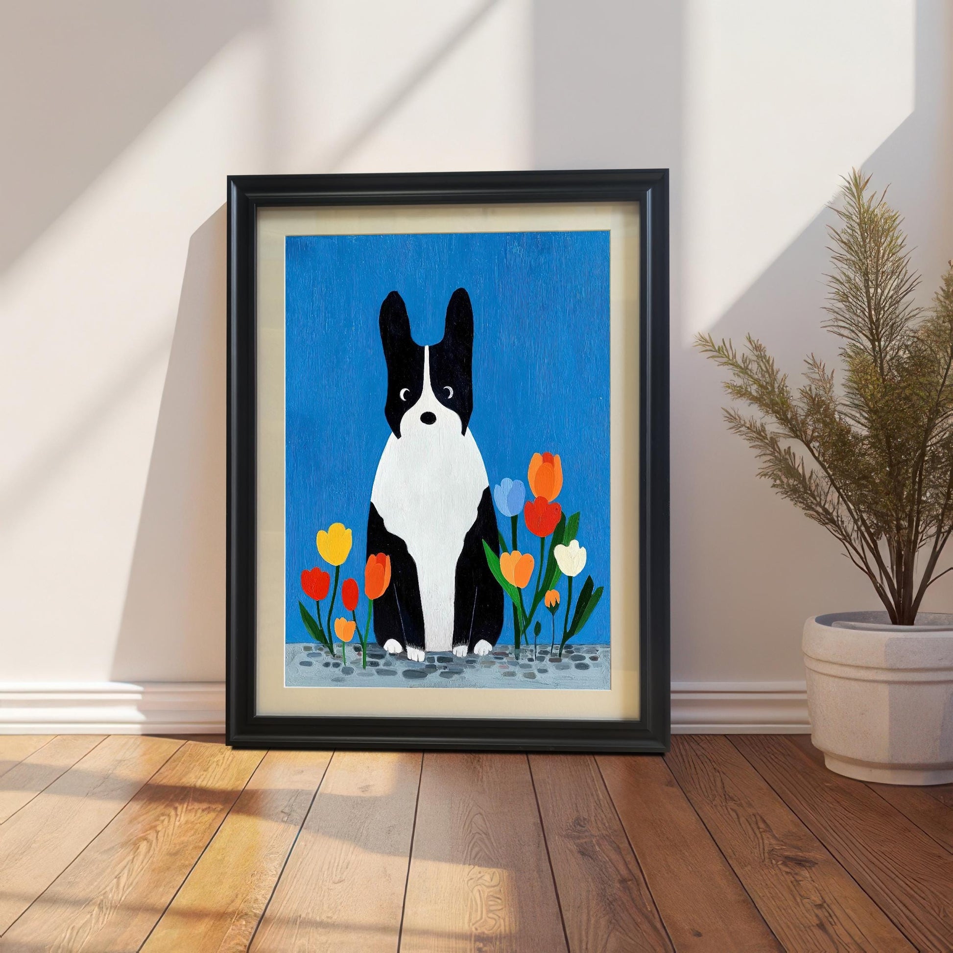 Framed art print of a black and white dog sitting among colorful tulips against a vibrant blue background. The print is in a black frame with a light-colored mat/mount, resting on a wooden floor against a white wall with sunlight streaming across it, next to a potted plant.