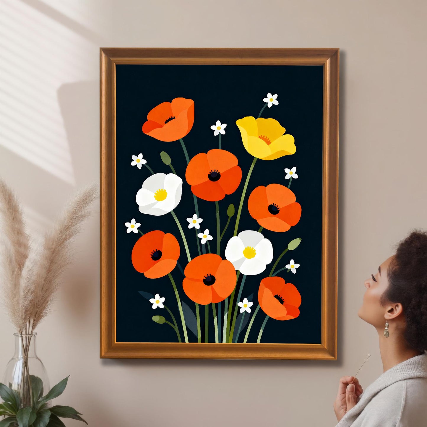 A framed art print of colorful, modern poppies with a dark background is hanging on a light beige wall. The print is in a gold-colored frame. A woman is visible on the right, looking up at the art. Dried reeds in a vase are on the left.
