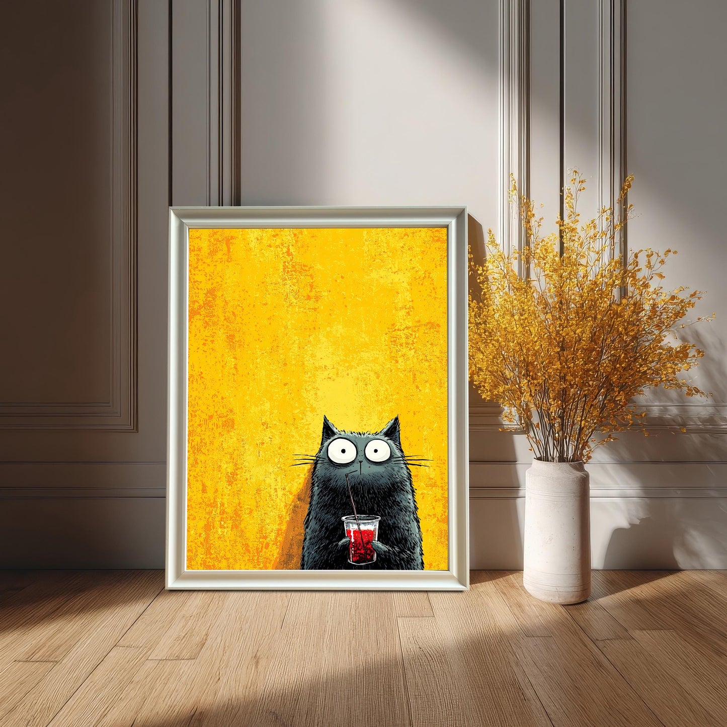 A framed art print featuring a cartoonish black cat with wide eyes drinking a red beverage through a straw, set against a bright yellow, textured background. The print is in a white or silver-colored frame, leaning on a hardwood floor against a white wainscoted wall. A tall, cream-colored vase with dried yellow flowers sits next to it.