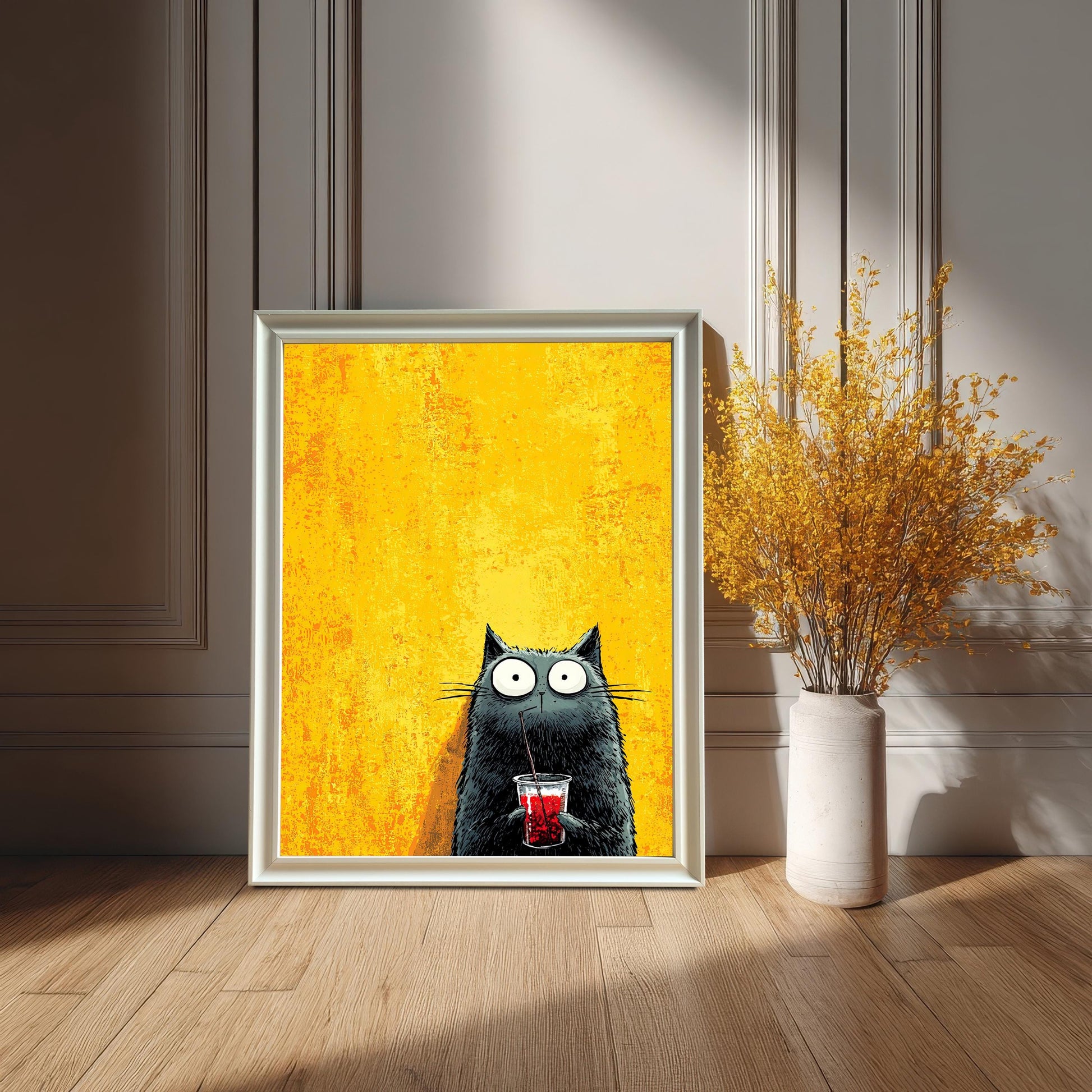A framed art print featuring a cartoonish black cat with wide eyes drinking a red beverage through a straw, set against a bright yellow, textured background. The print is in a white or silver-colored frame, leaning on a hardwood floor against a white wainscoted wall. A tall, cream-colored vase with dried yellow flowers sits next to it.