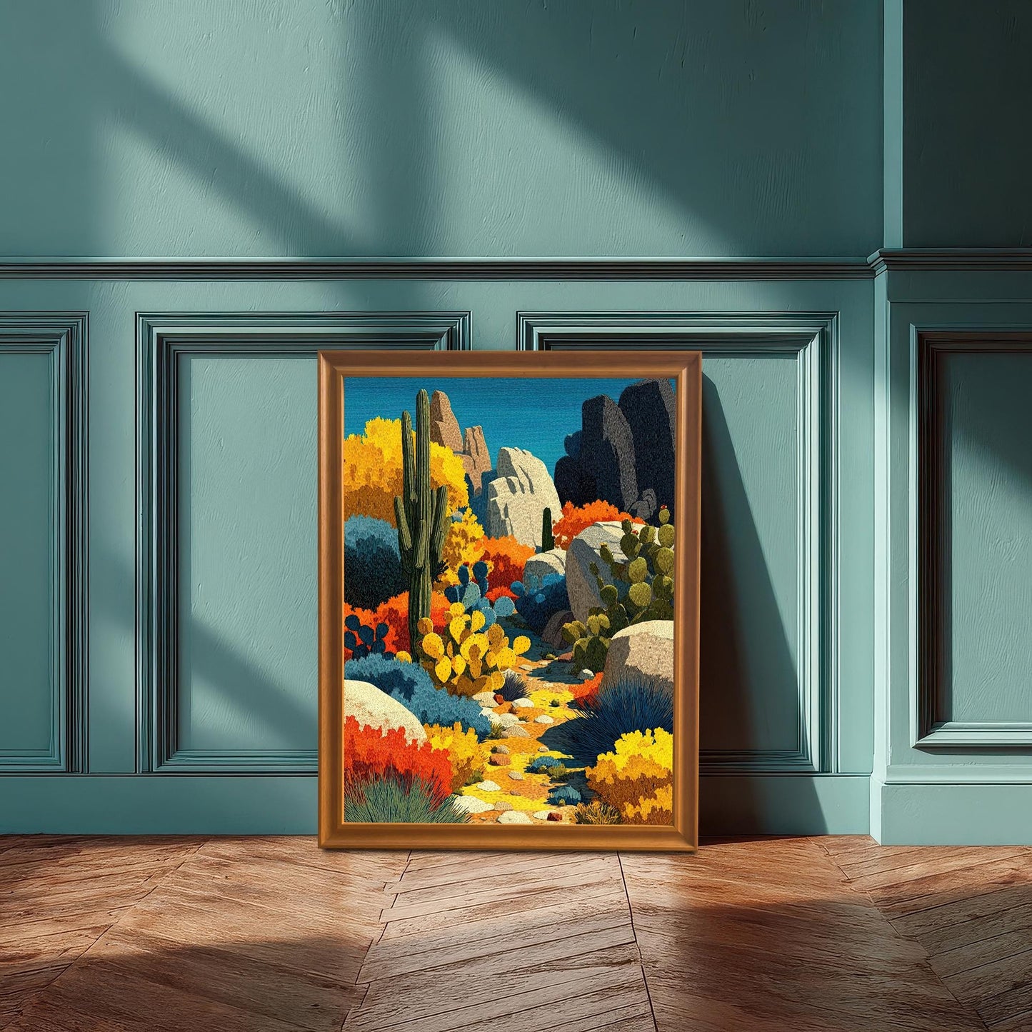A framed, colorful abstract desert landscape print leaning against a teal wainscoting wall, with sunlight casting shadows on the floor and wall. The frame is a warm, orange-gold wood.