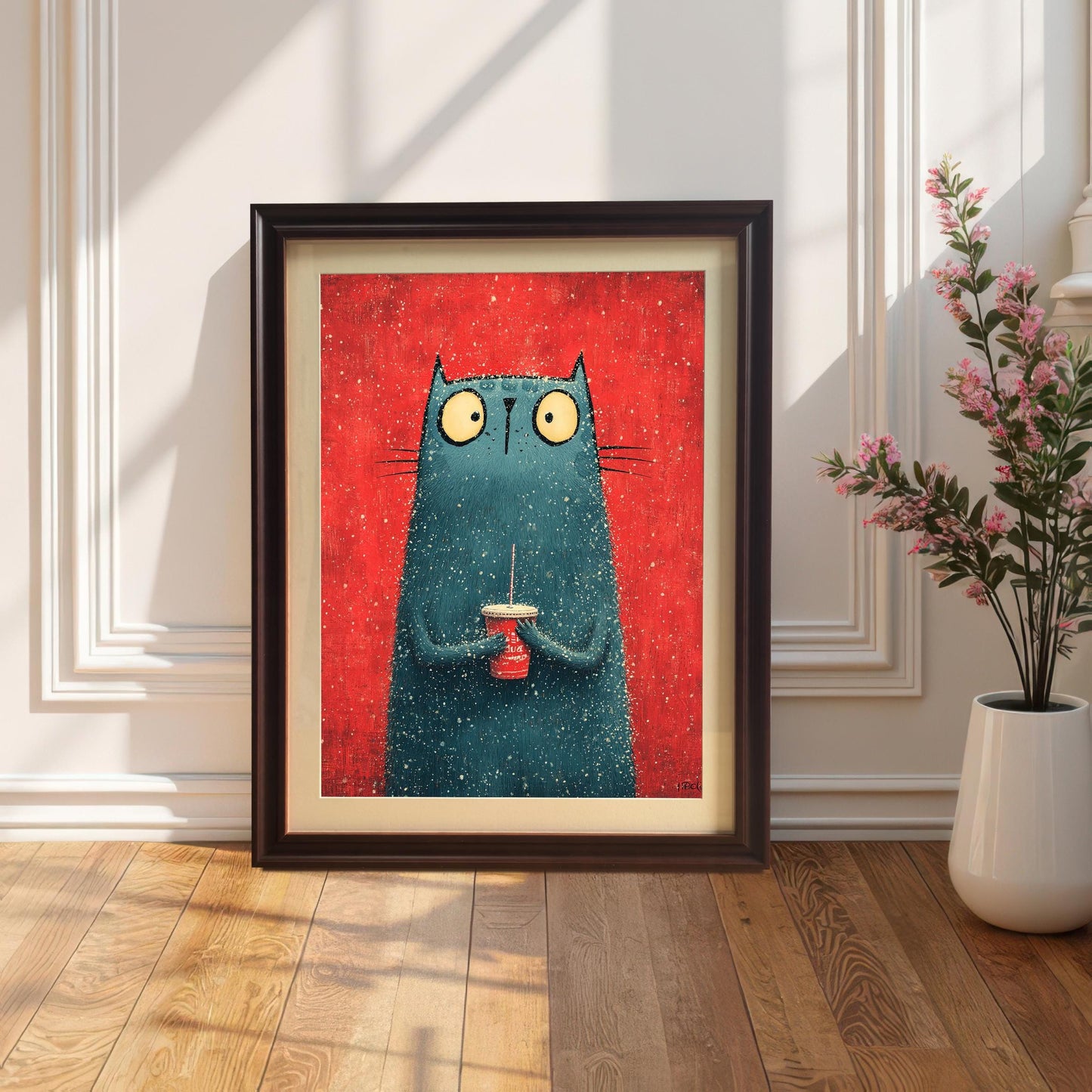 A framed art print featuring a stylized, fuzzy, blue-green cat holding a red cup against a red background. The print is in a Coffee Frame (dark brown frame with a light mount/border) and is leaning on a hardwood floor against a white, wainscoted wall. A white vase with pink flowers is on the right.