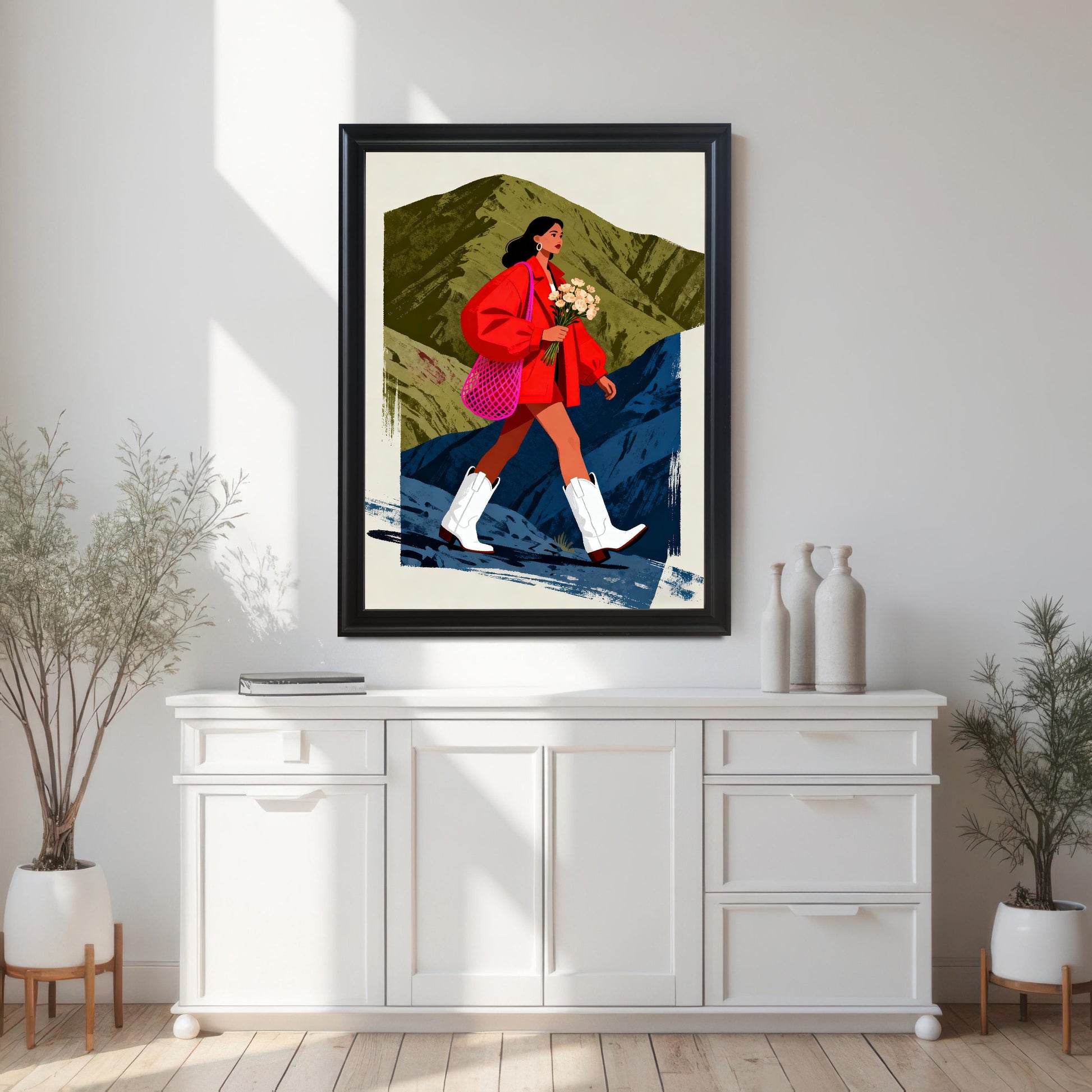 A framed art print featuring a stylized illustration of a woman in a red jacket and white cowboy boots walking across a mountainous landscape with olive green and deep blue brushstrokes. The print is in a Black Frame and is centered above a long, white console table.